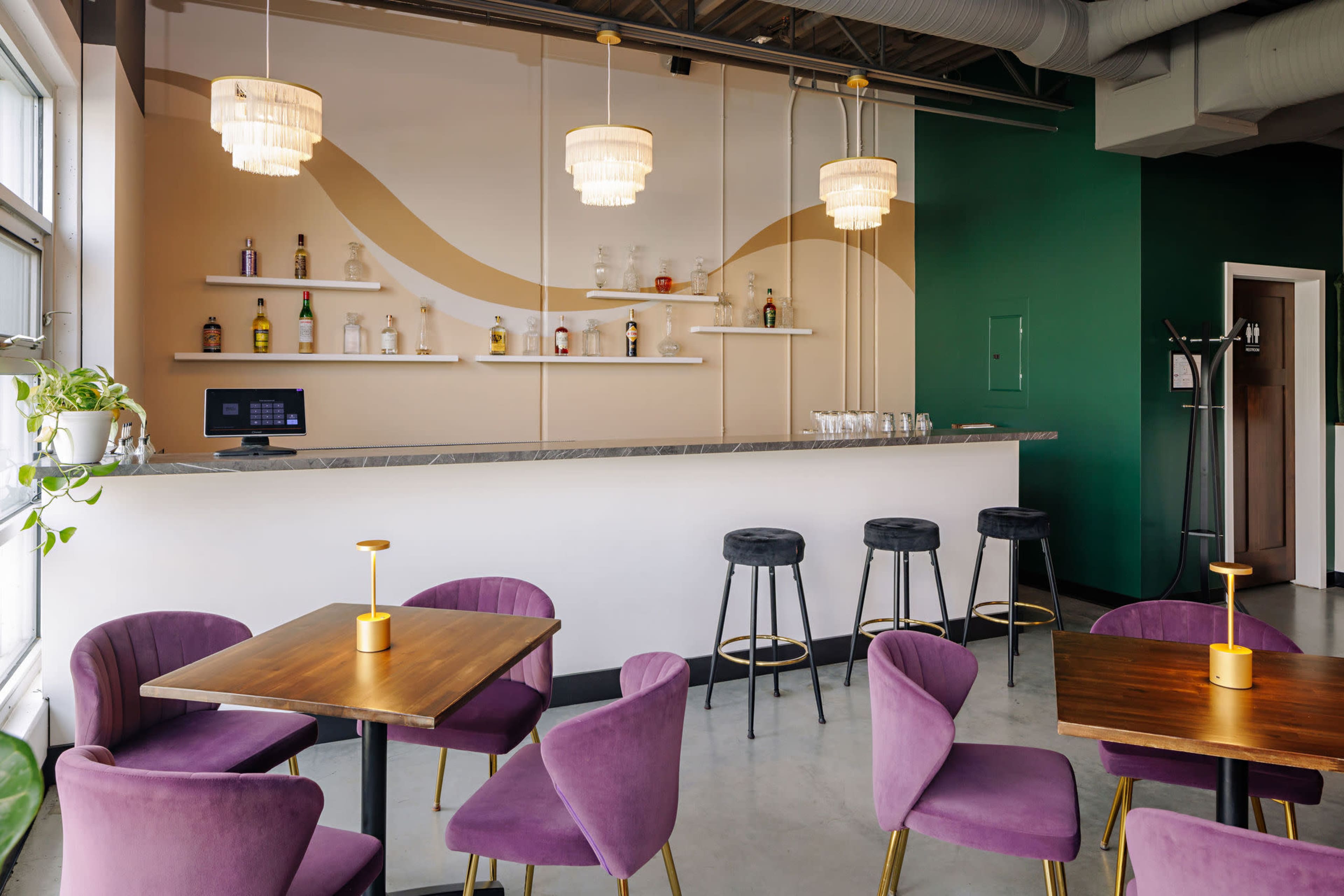 The image shows a modern bar area with a light-colored counter, shelves displaying bottles, and purple chairs around wooden tables.