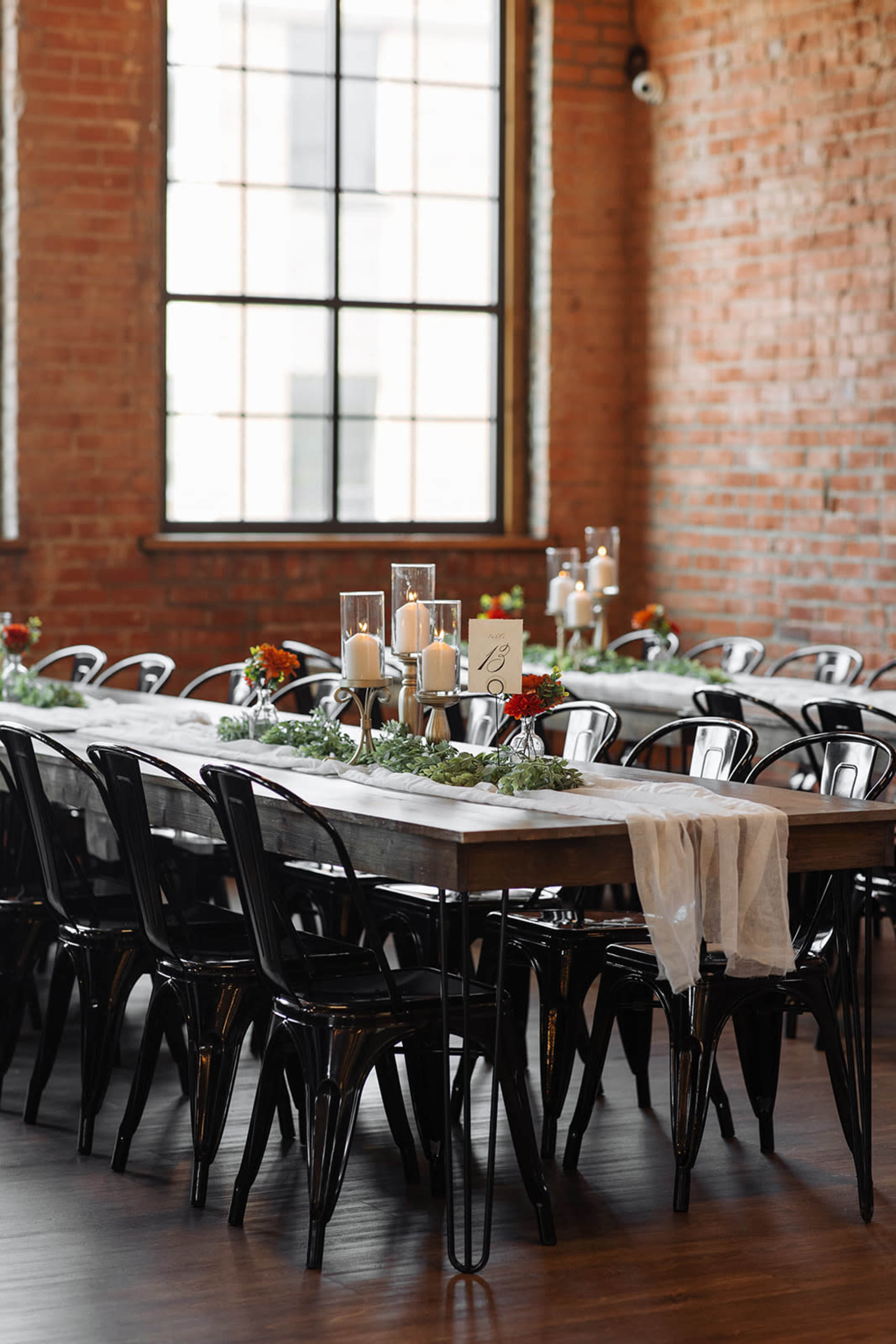 A long wooden dining table is set with white table runners and surrounded by black metal chairs in a room with exposed brick walls and large windows.