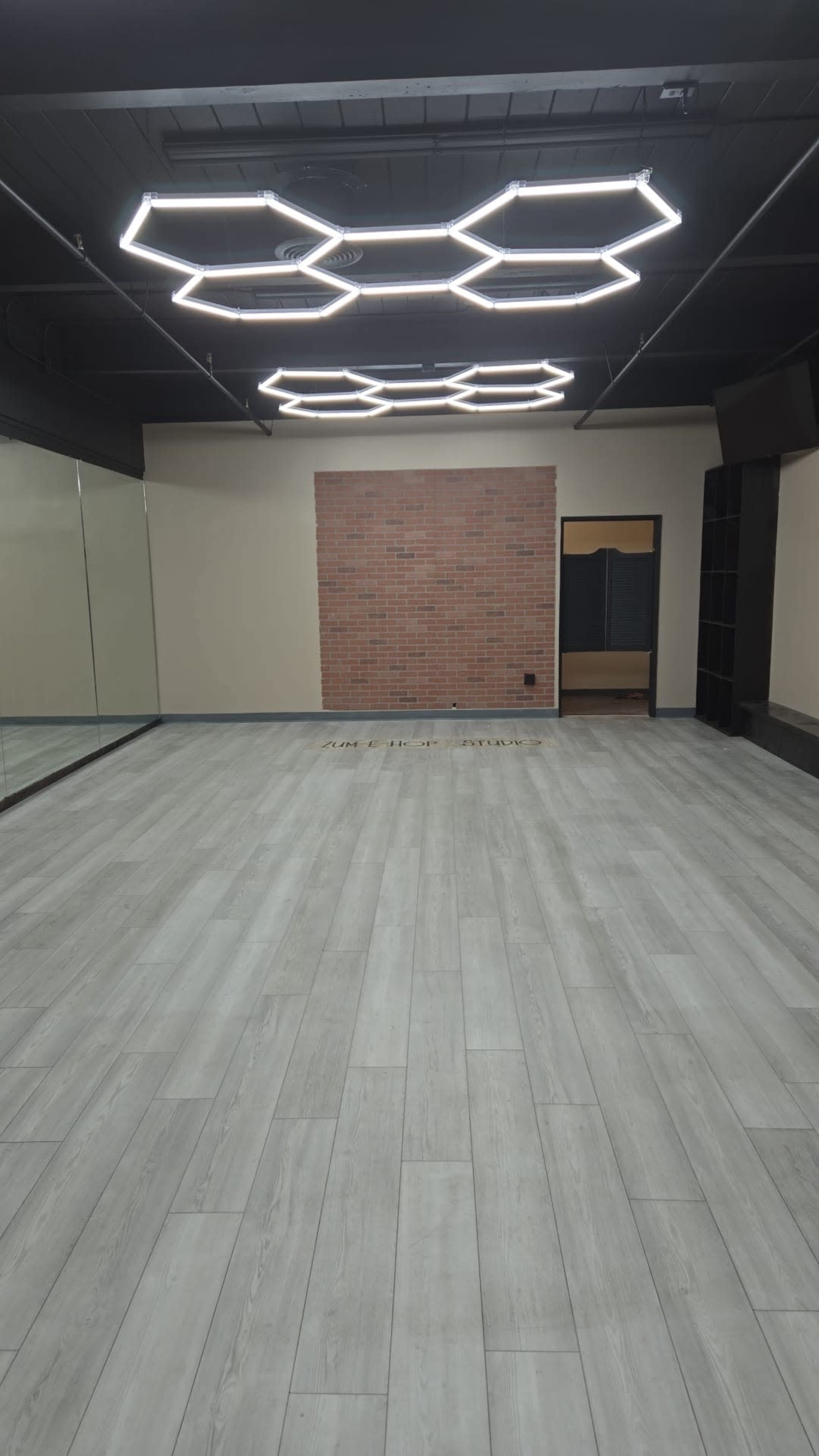The image shows an empty studio space with a wooden floor, a brick accent wall, and modern hexagonal light fixtures hanging from the ceiling.