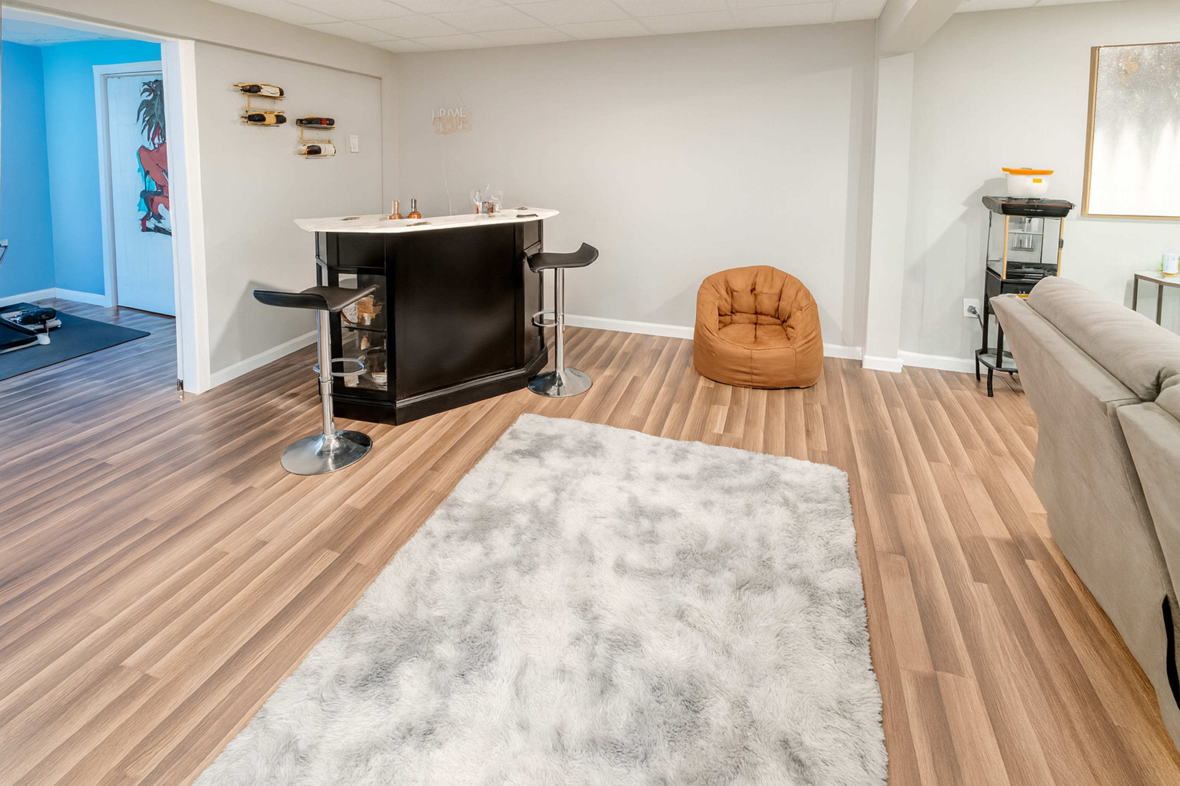The image shows a modern and spacious living area with light wood flooring, a bar counter with stools, a bean bag chair, and a plush area rug.