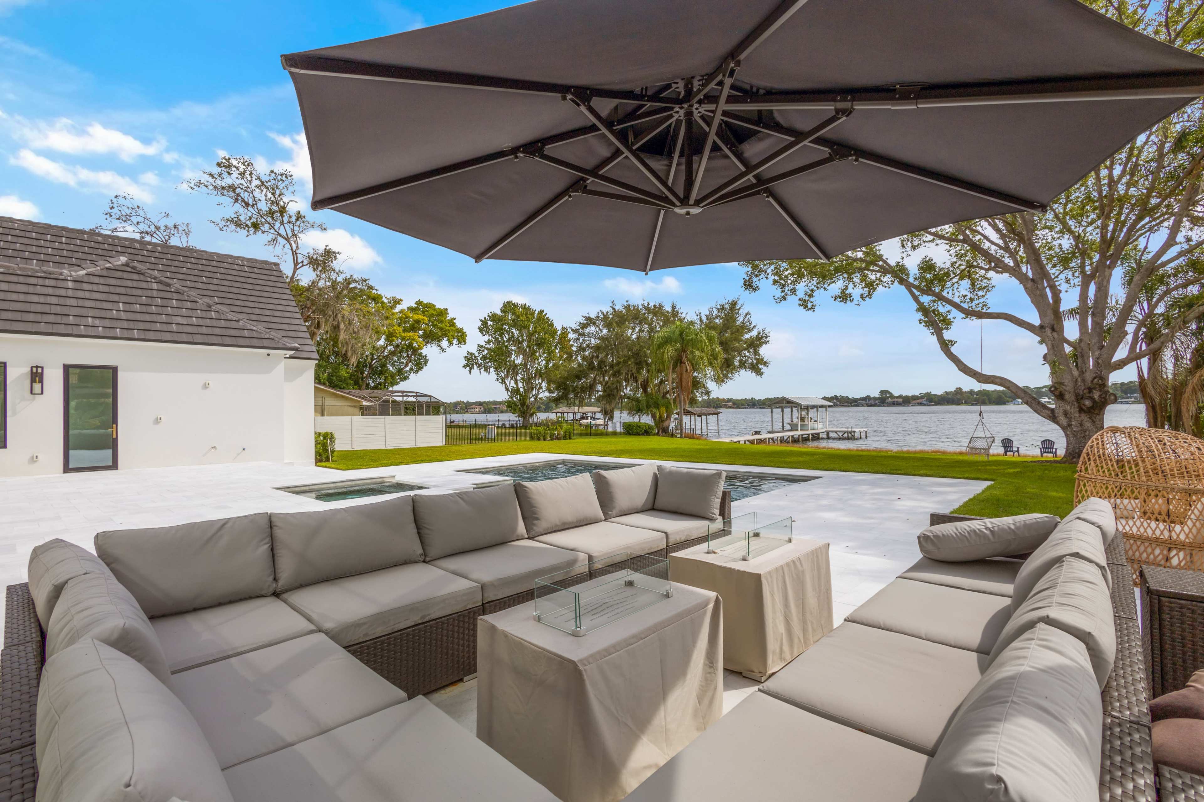 A modern outdoor seating area with a large umbrella overlooks a calm body of water surrounded by trees.
