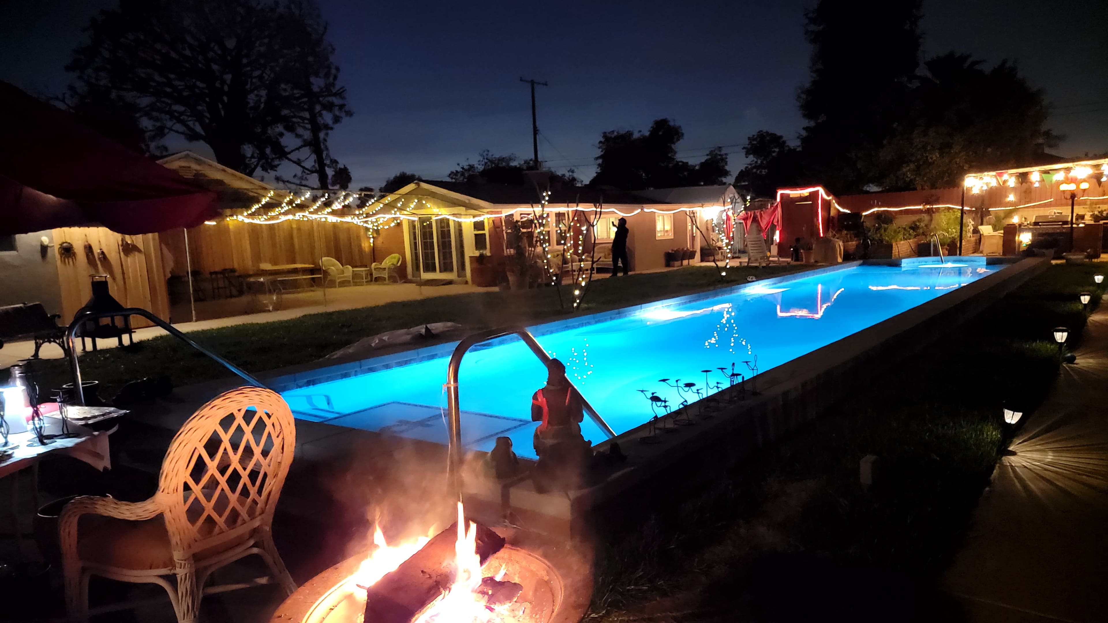 The scene features a brightly lit swimming pool surrounded by a backyard adorned with festive lights and a fire pit.