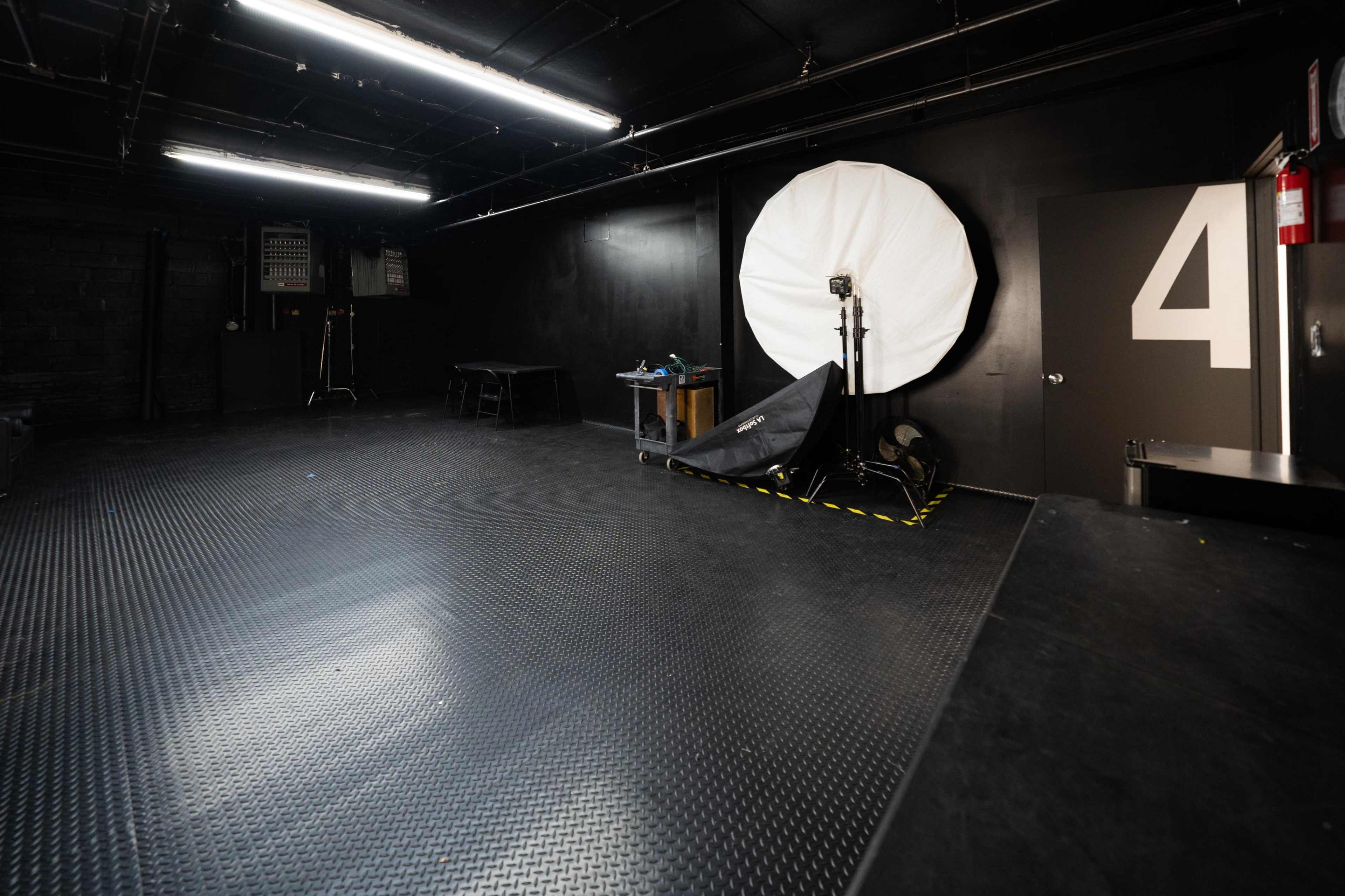 A photography studio with a large white umbrella softbox, a door marked with the number 4, and equipment on a table in the corner.