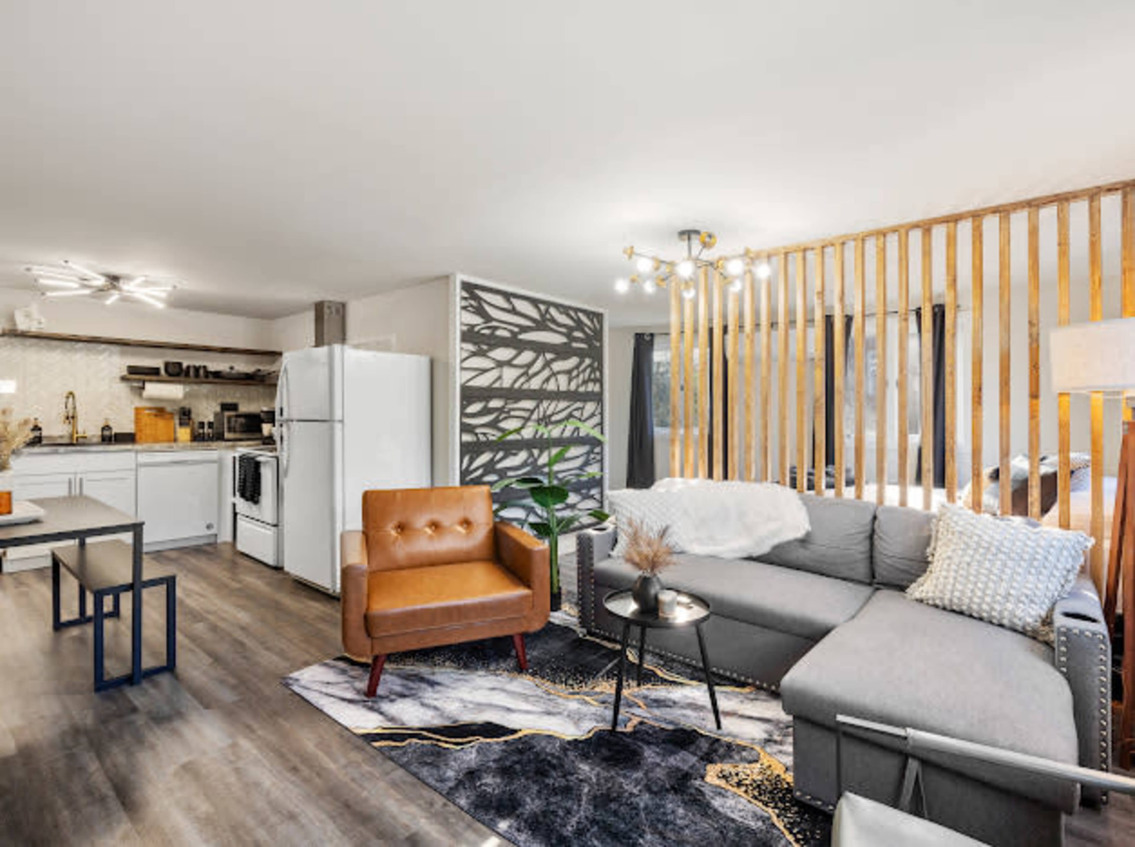 A modern living space featuring a gray sectional sofa, a brown leather armchair, a patterned rug, and a kitchen area with white cabinets and appliances.
