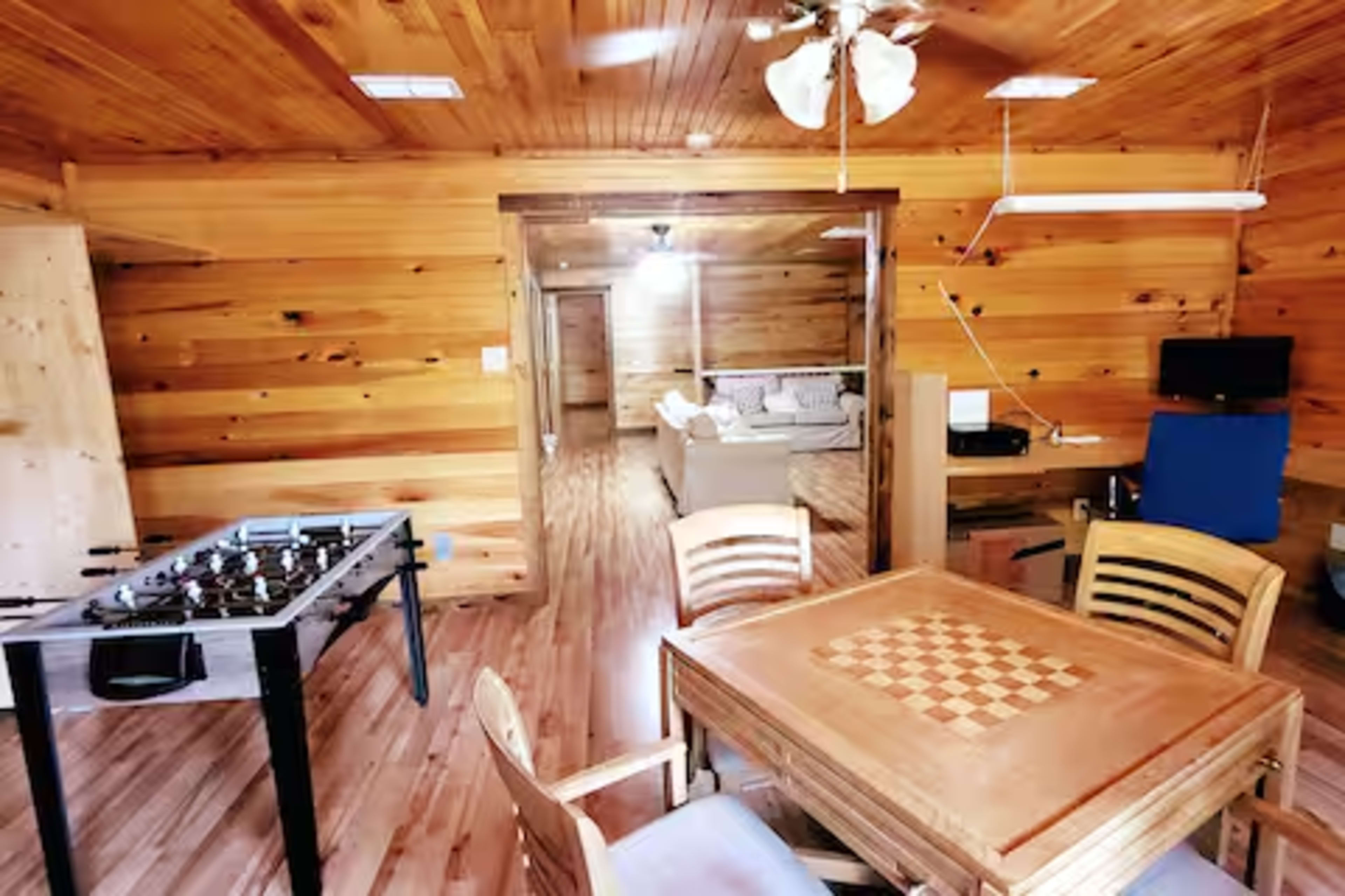 The image shows a wooden-paneled interior room featuring a foosball table, a dining table with a checkered pattern, and a seating area with a couch.