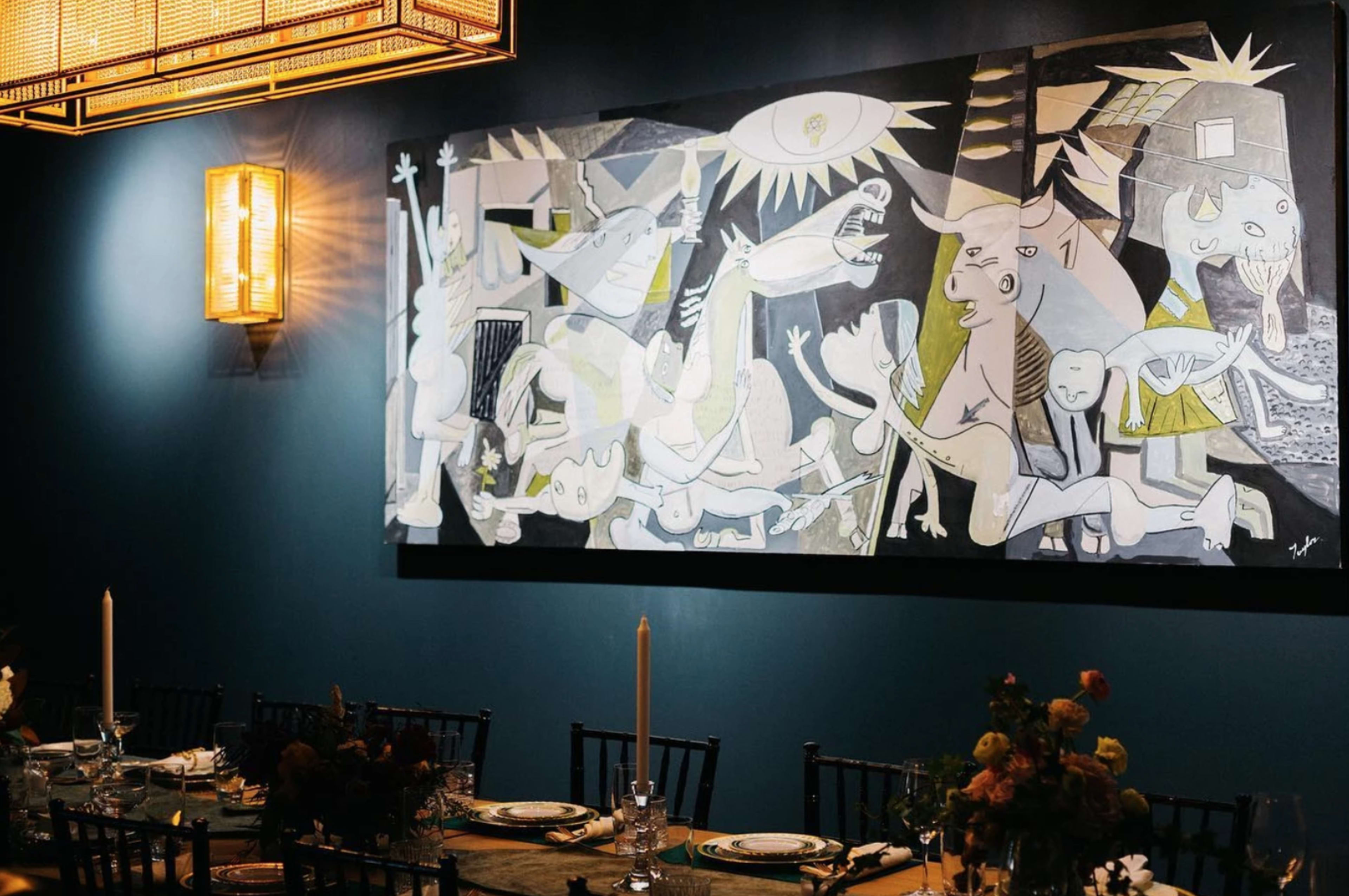 A large abstract painting hangs on a dark blue wall above a long dining table set with candles and floral arrangements.