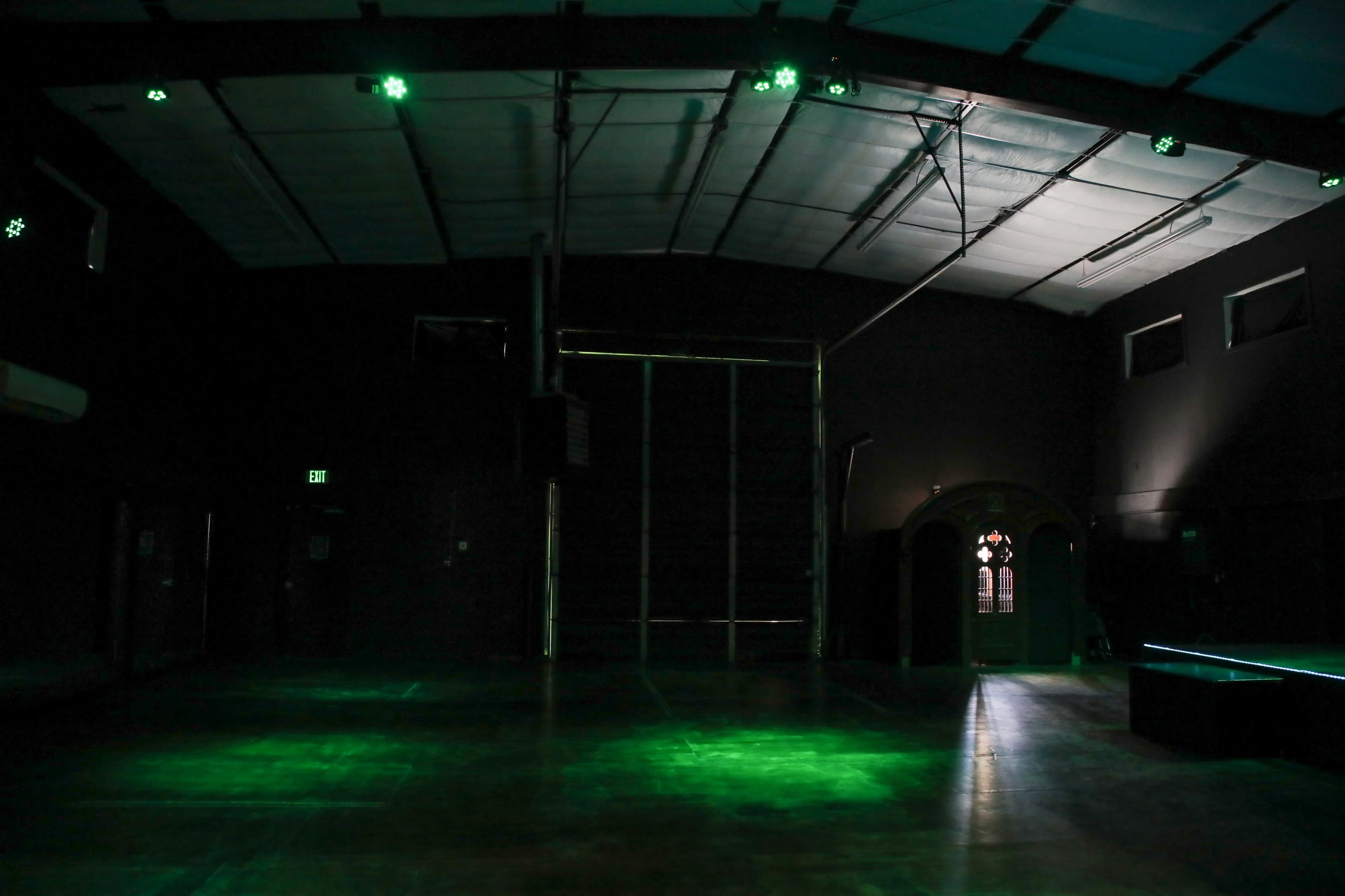 The image shows a dimly lit, empty room with a high ceiling, featuring green lights illuminating the floor and a large door at the far end.