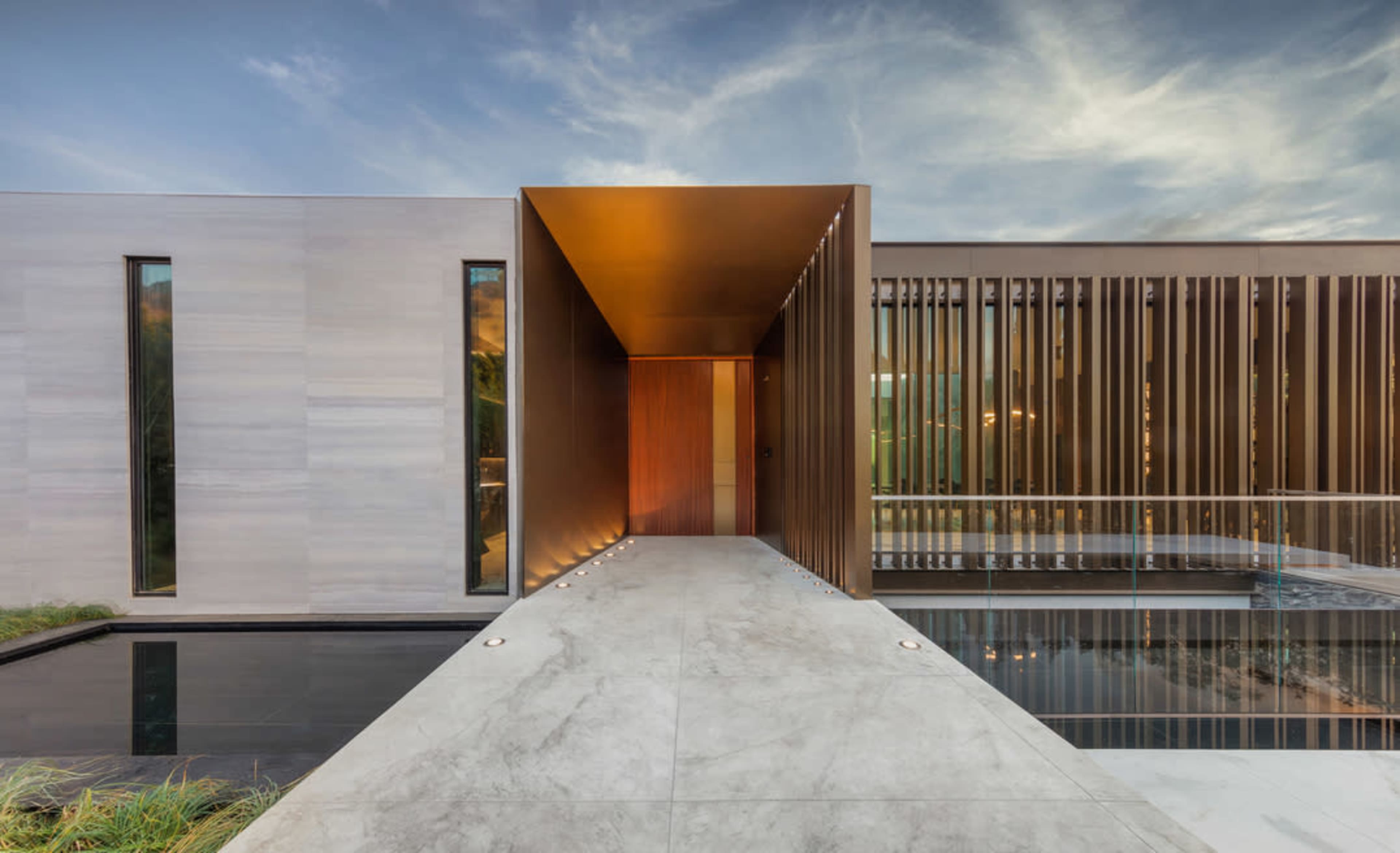 A modern architectural entrance to a sleek house, featuring a pathway that leads to a large door, bordered by large windows and a reflecting pool.