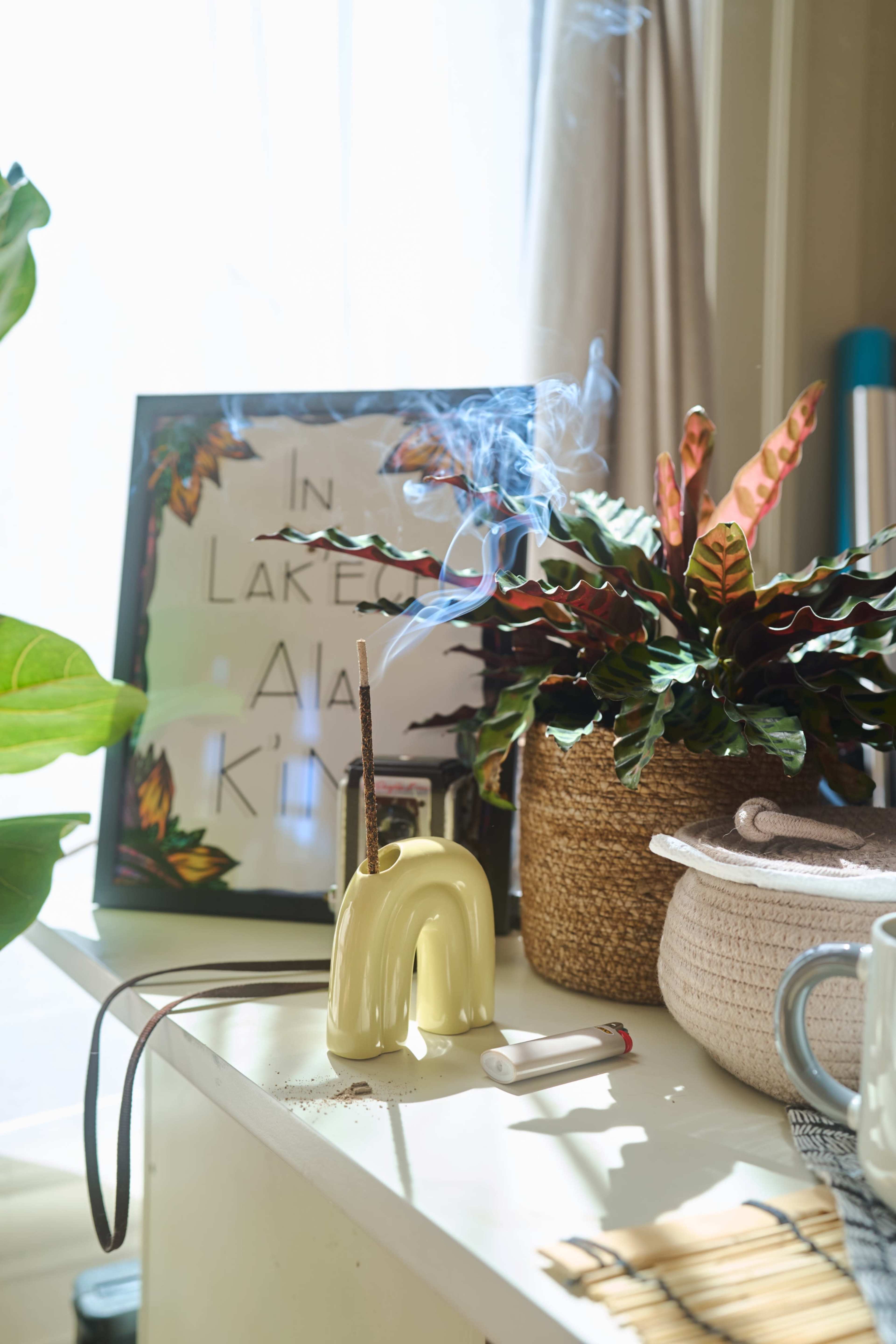 A small, yellow incense holder with incense sticks rests on a table beside a potted plant and a framed picture.