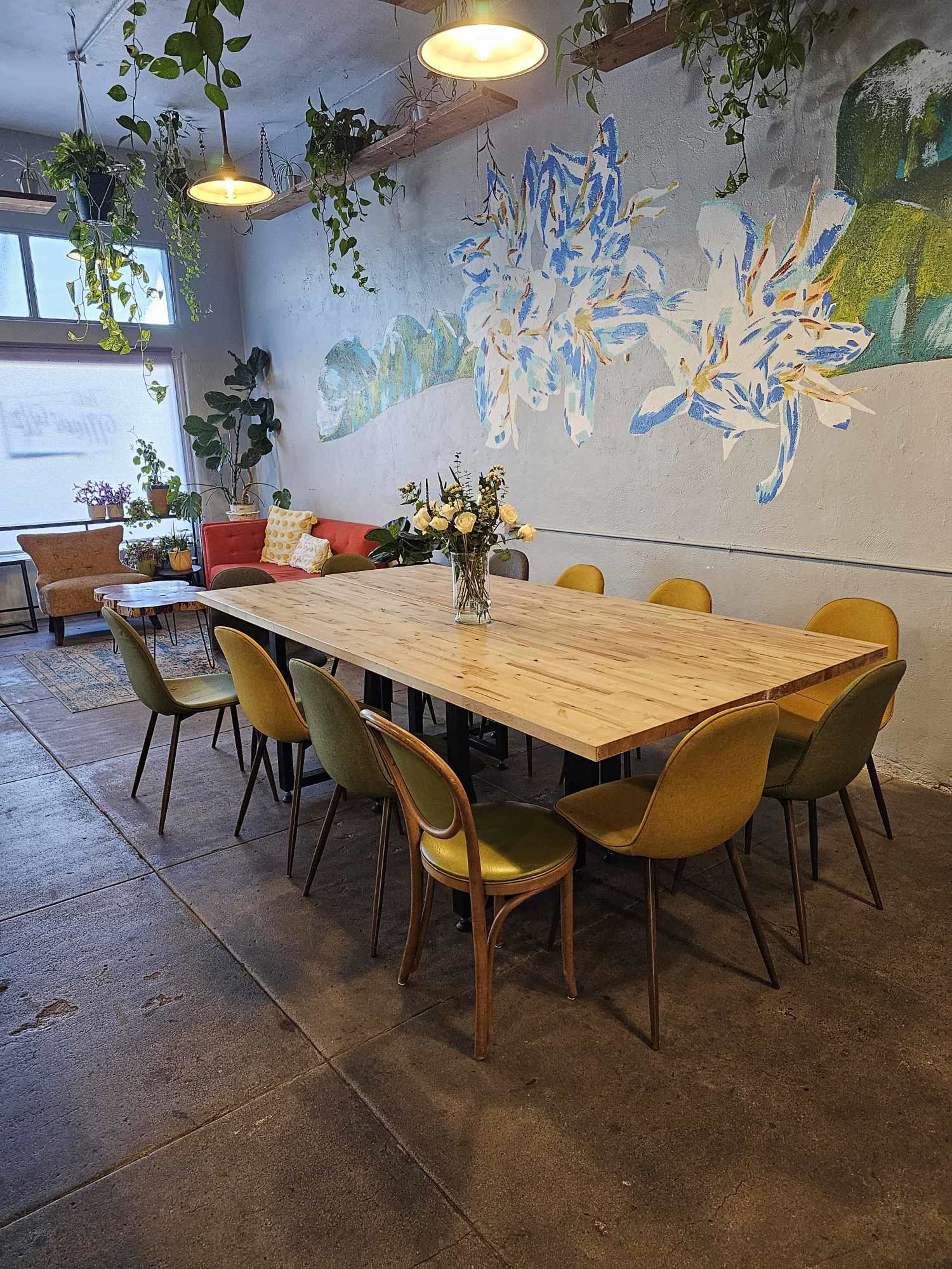 A large wooden dining table surrounded by green and yellow chairs in a room decorated with plants and a mural of flowers on the wall.