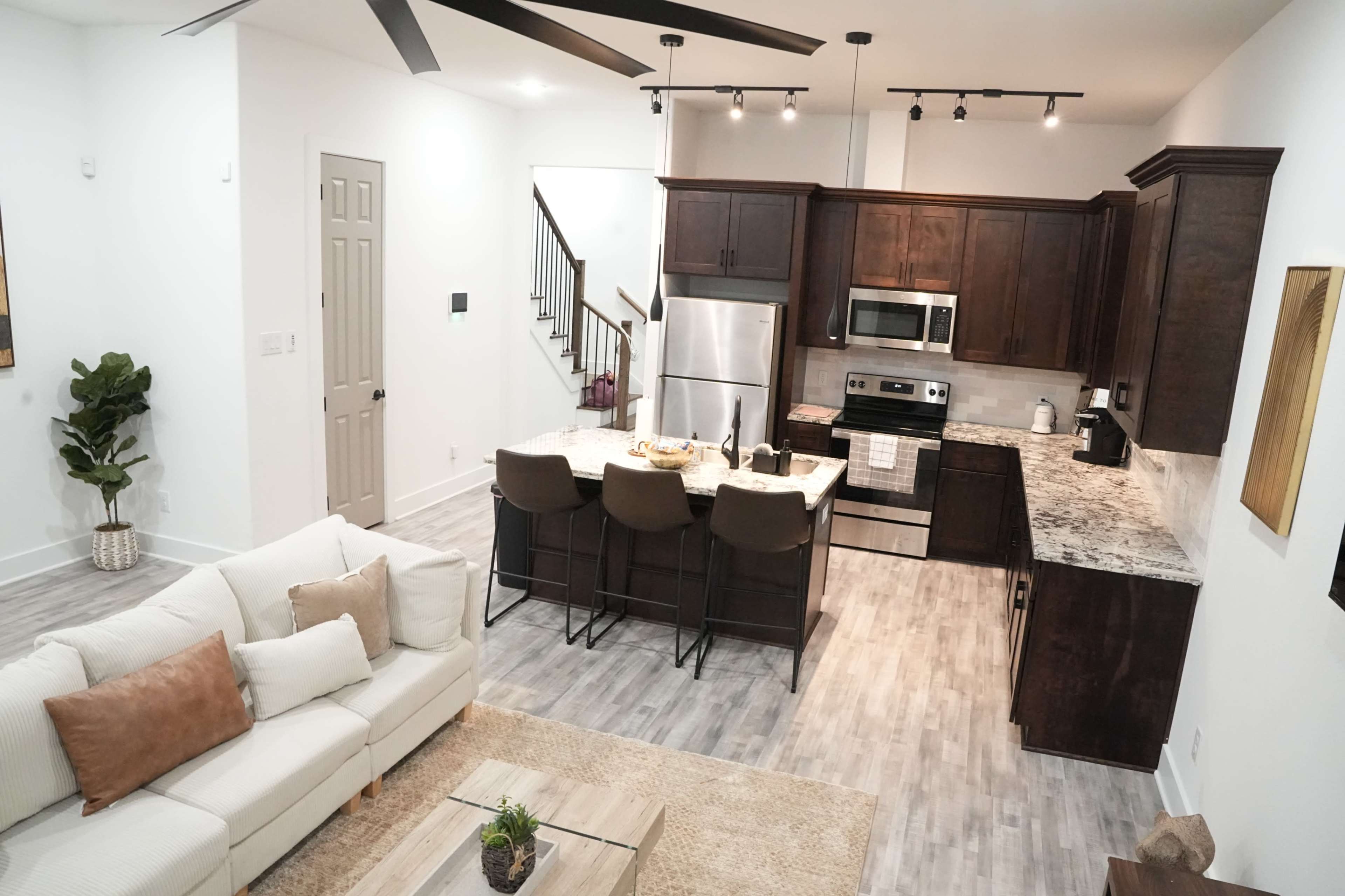 The image shows a modern open-concept kitchen and living area with a light-colored couch, a kitchen island with bar stools, and stainless steel appliances.