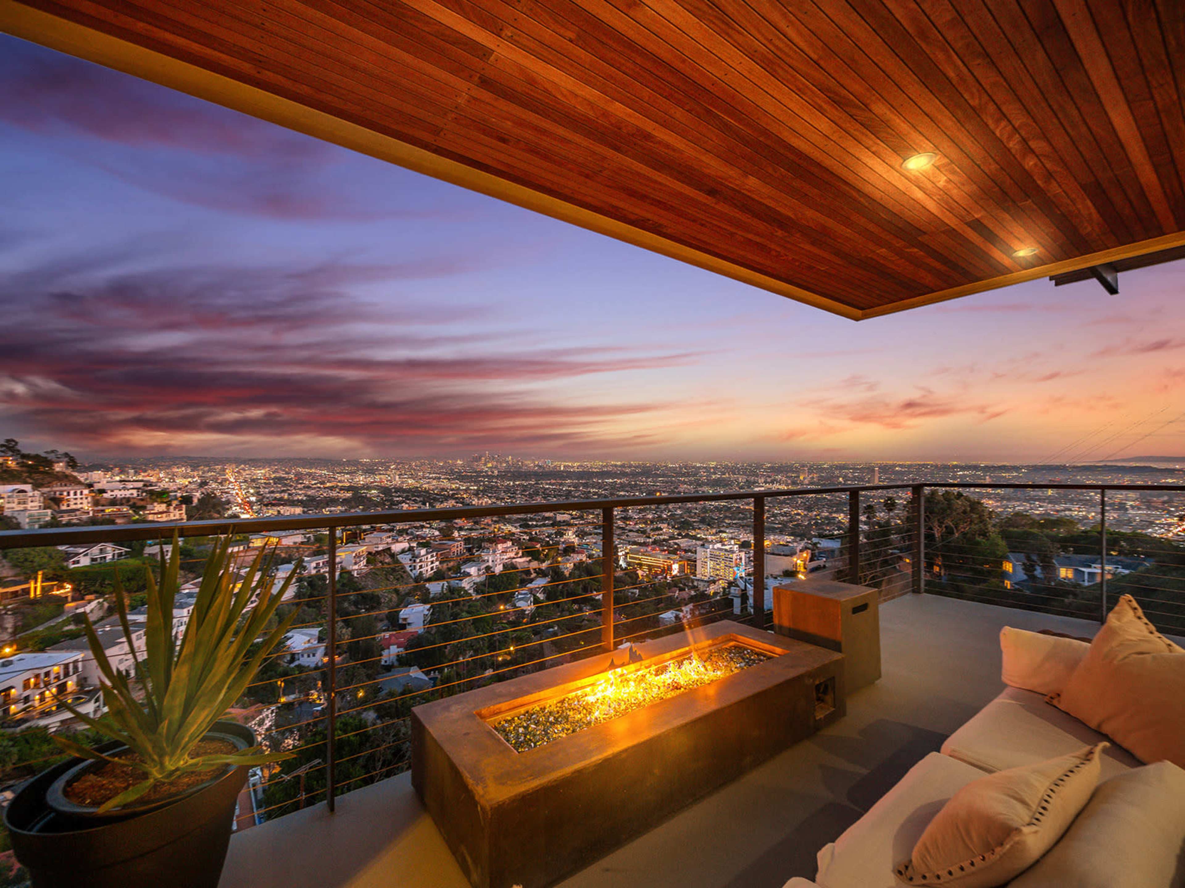 A modern balcony features a fire pit overlooking a city skyline at sunset, with expansive views of the illuminated buildings below.