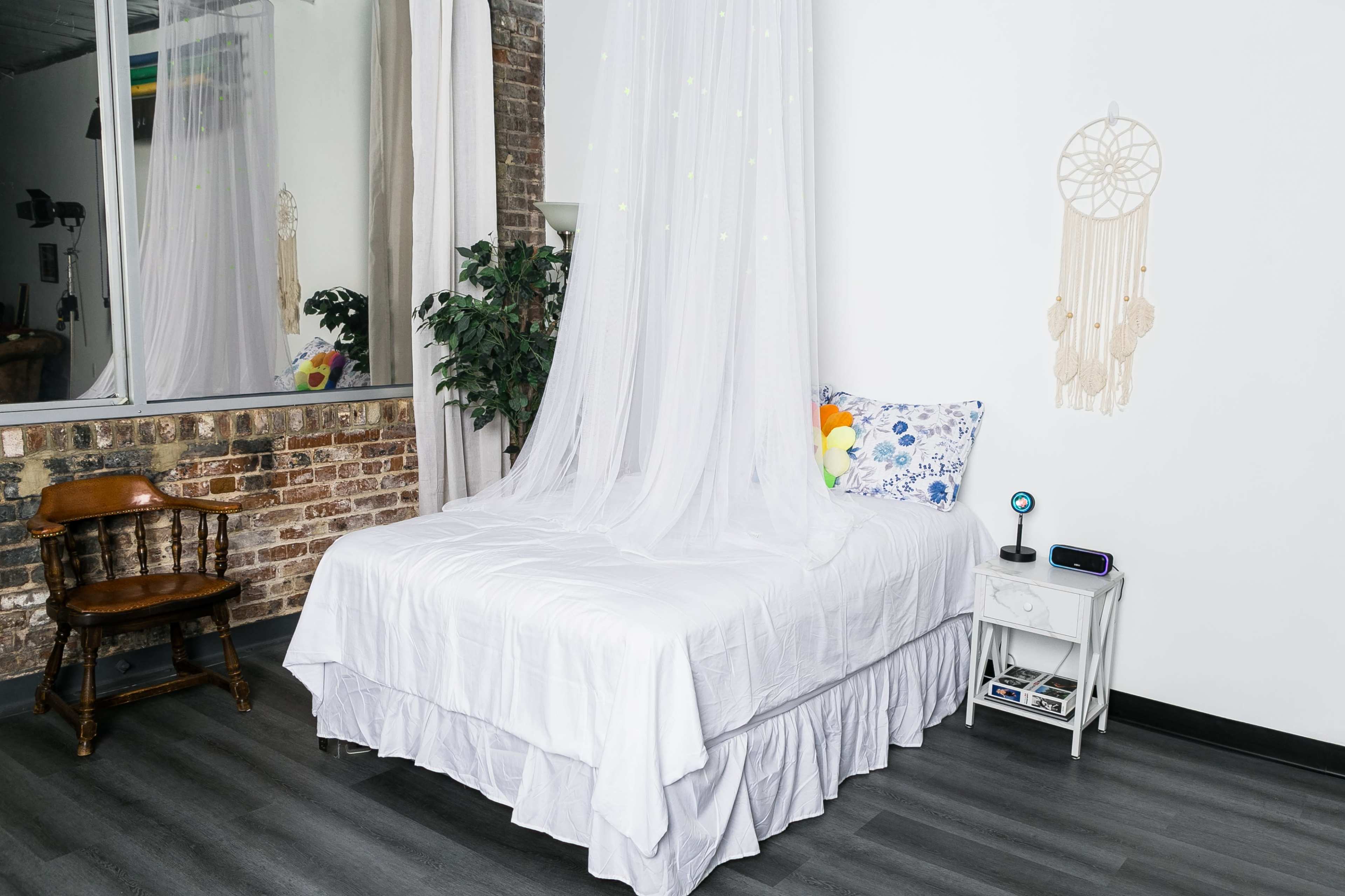 The image shows a bedroom with a white bed covered by a sheer canopy, a wooden chair, and decorative elements like a dreamcatcher and a nearby plant.