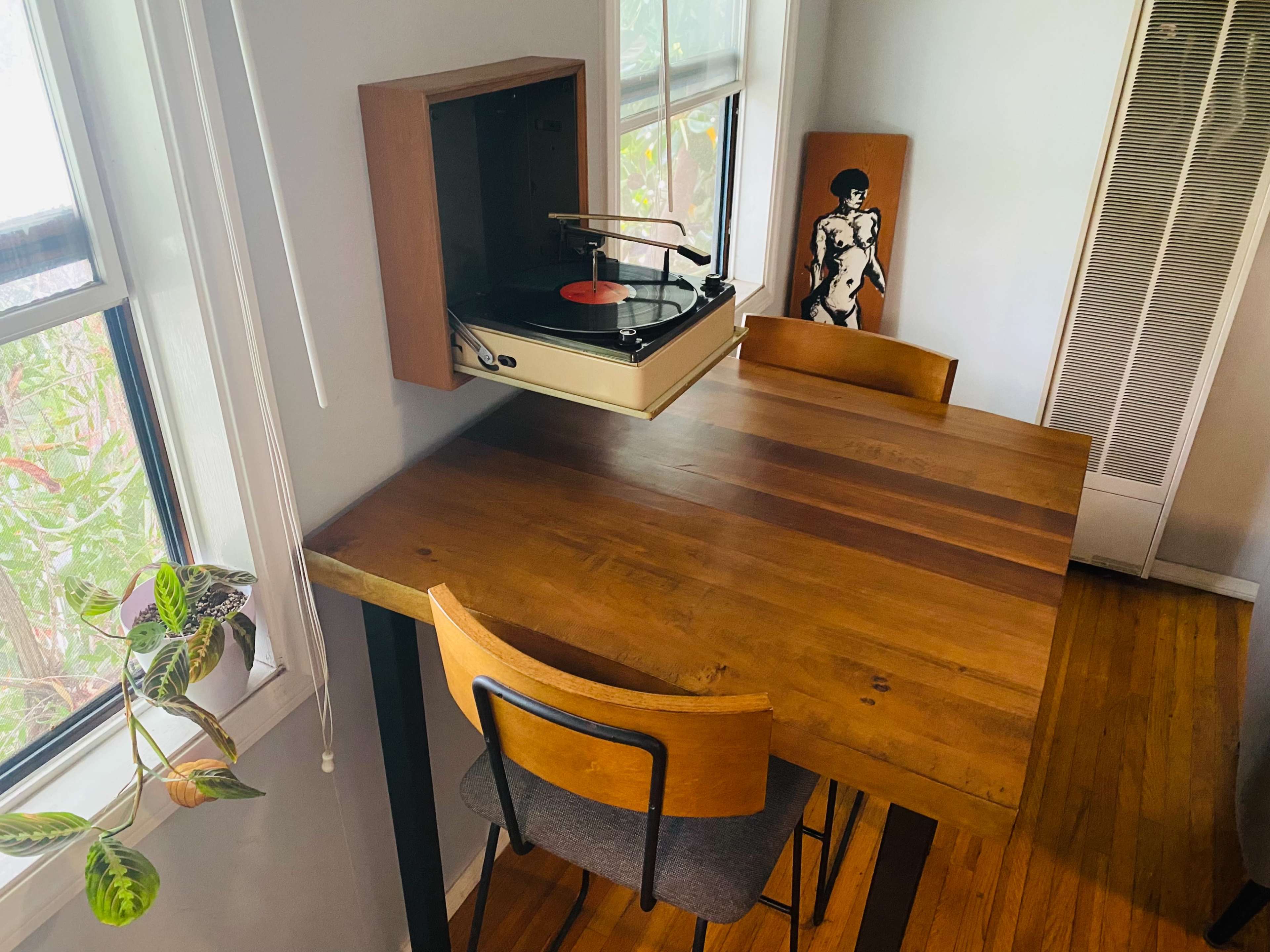 A wooden dining table with two chairs sits near a wall-mounted record player and a window, with a plant in the corner.