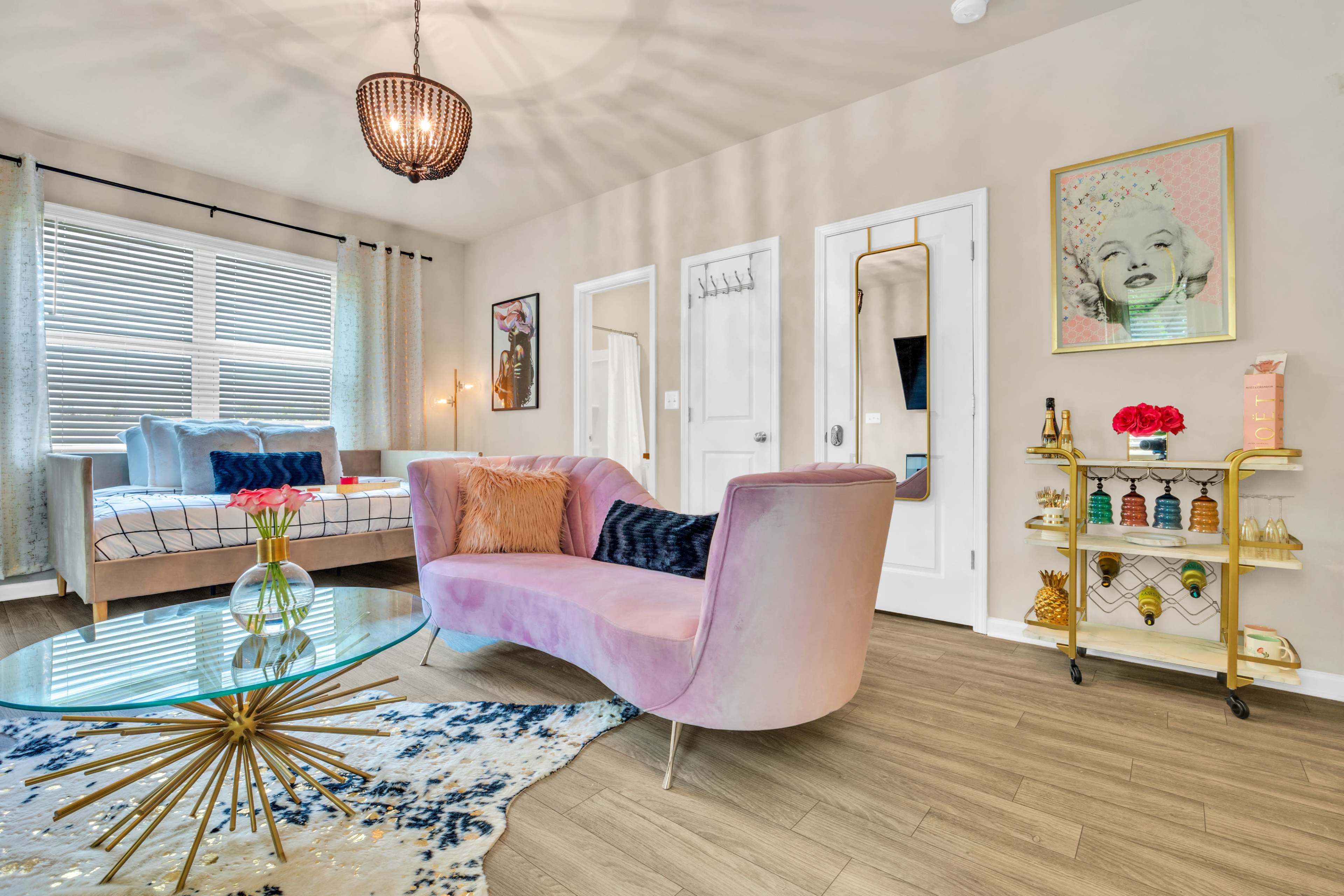 A modern living room featuring a pink curved sofa, a glass coffee table with a gold base, and a vintage-style bar cart, all arranged against a backdrop of light-colored walls and large windows.