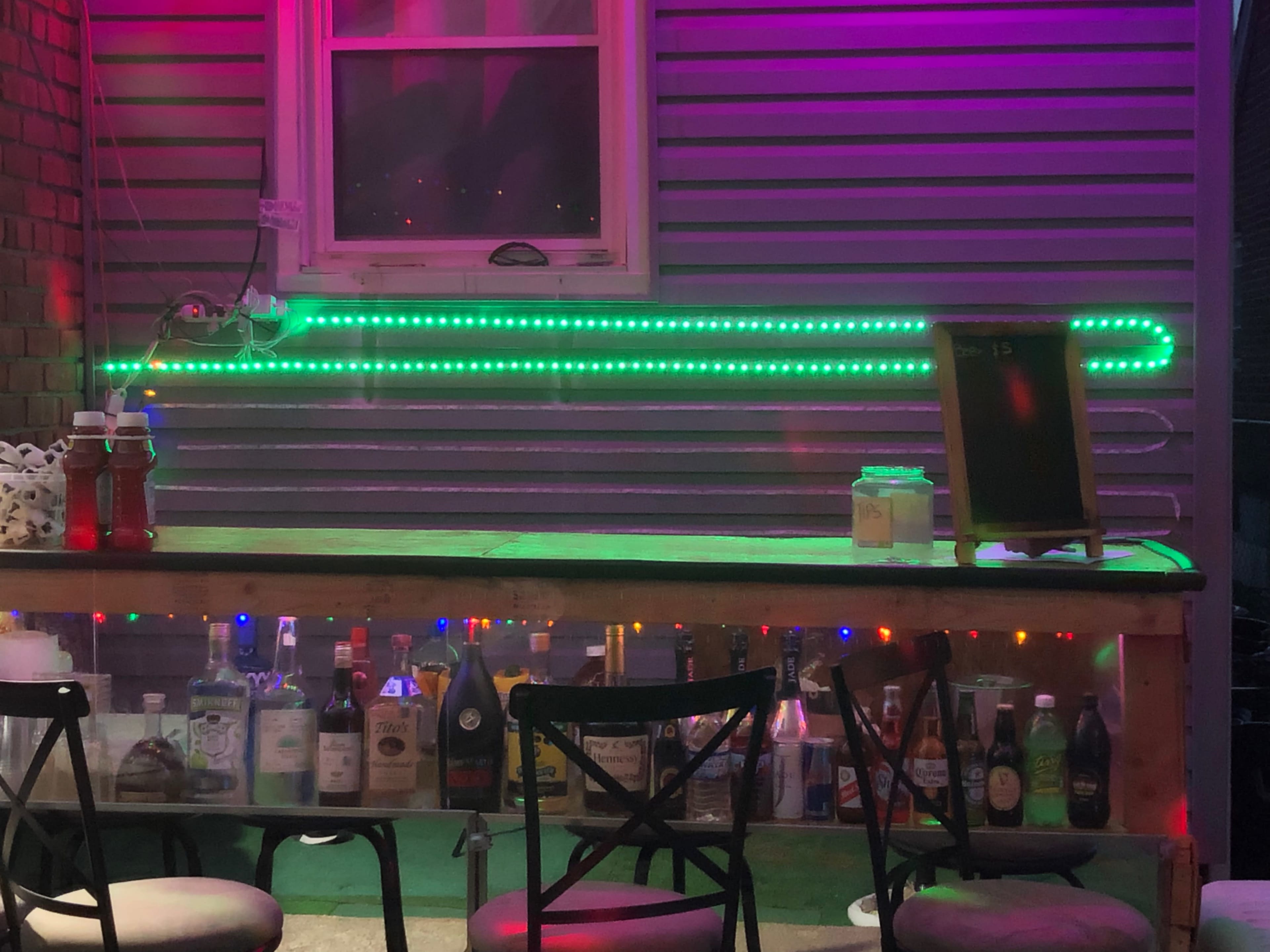 A bar area with a wooden counter, illuminated by green neon lights, showcases various bottles of liquor against a backdrop of a silver corrugated wall.