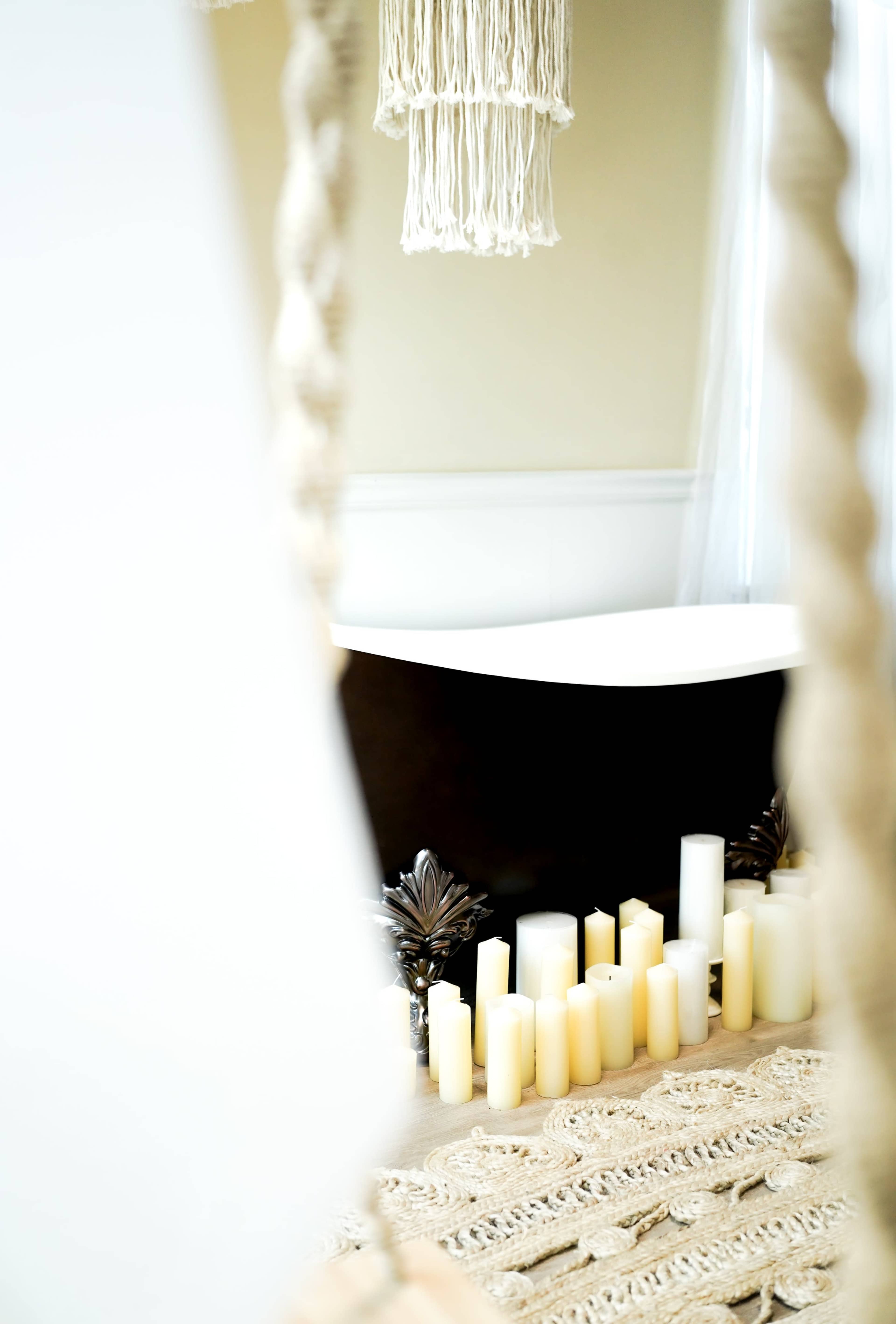 A freestanding bathtub is positioned against a wall, surrounded by various candles on a woven rug.