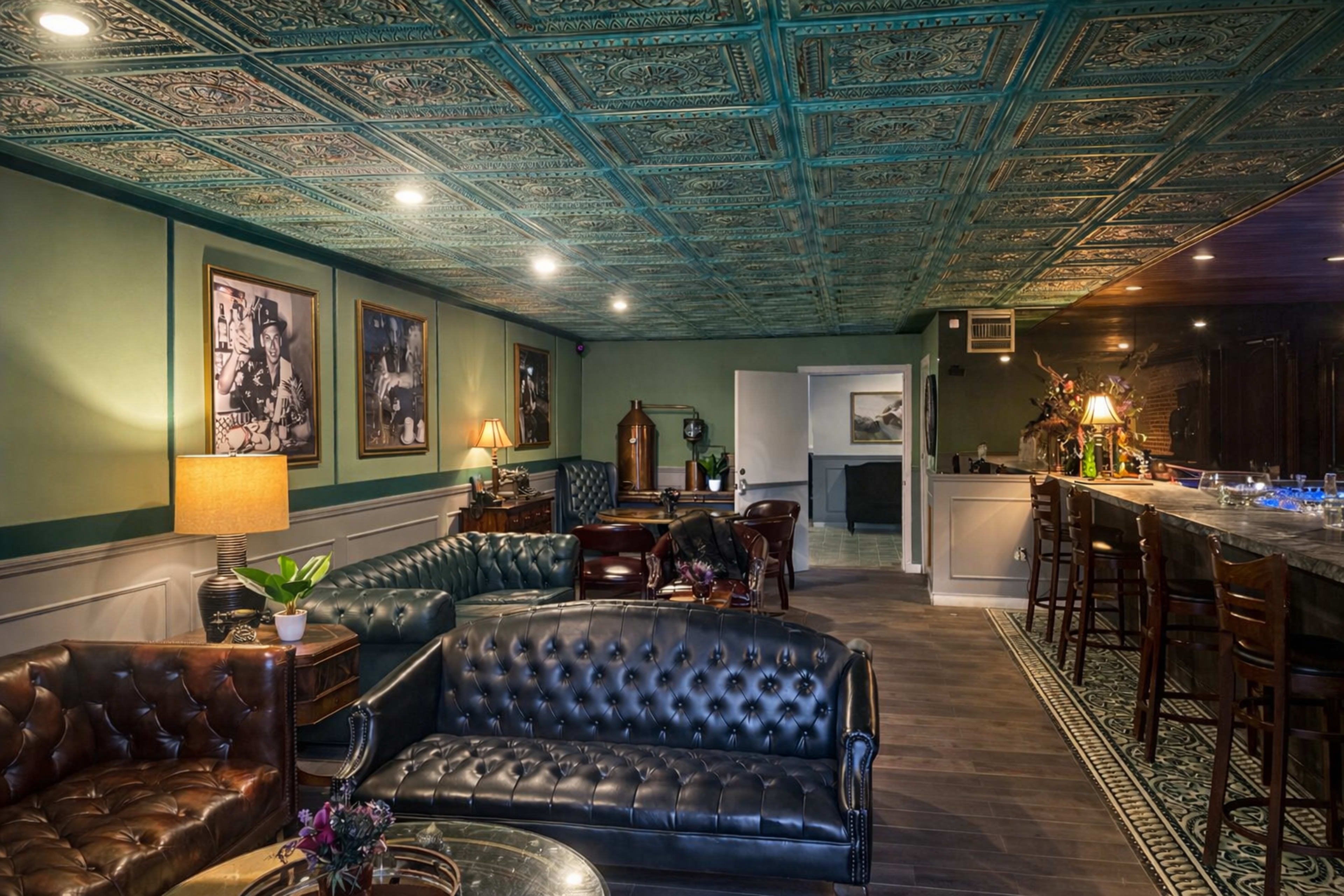 The image shows a spacious interior of a lounge featuring dark leather sofas, a bar area, and decorative ceiling tiles.