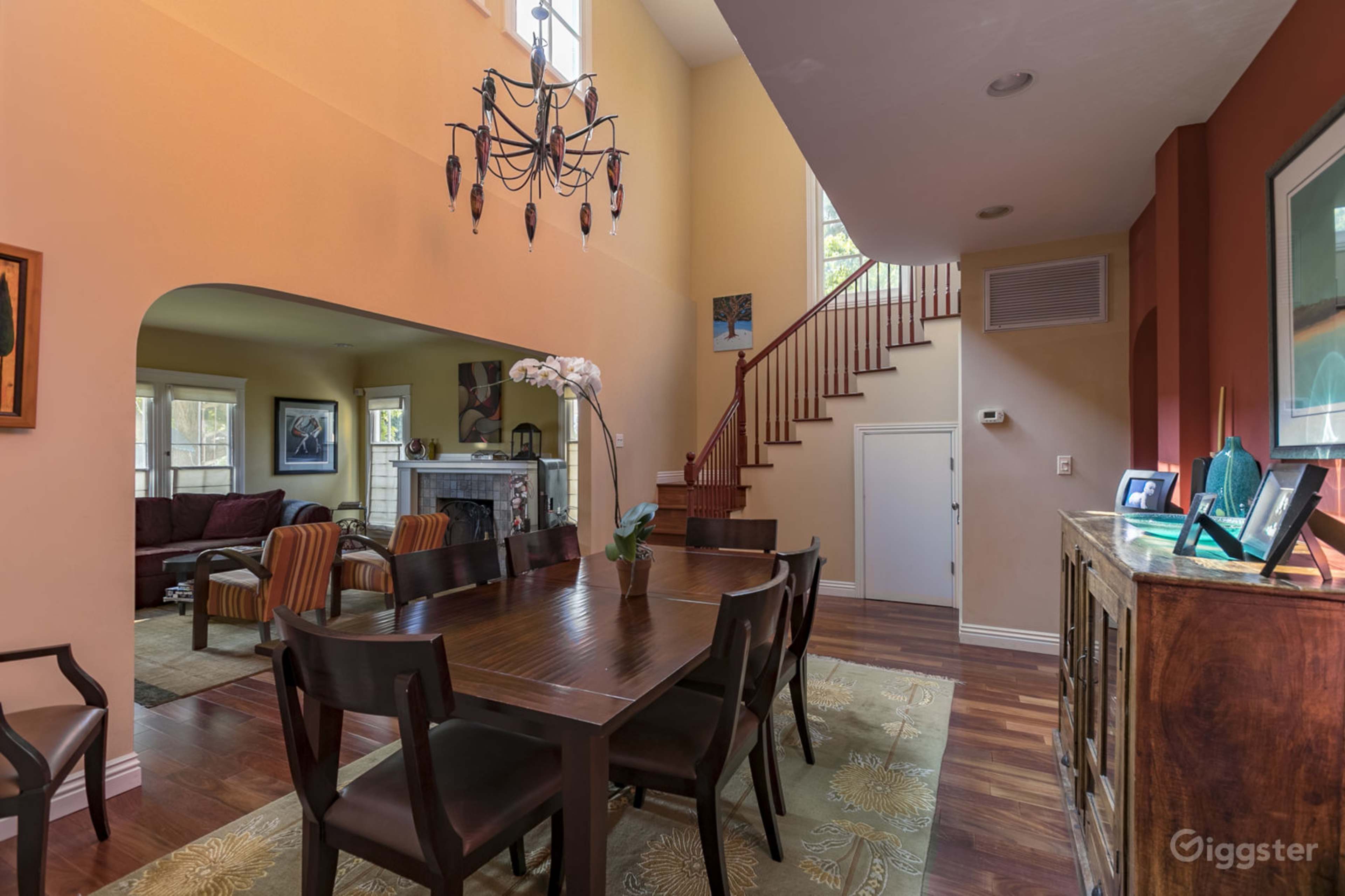 The image shows a dining area with a large table and chairs, adjacent to a staircase leading to the upper level, and features artwork and a chandelier.