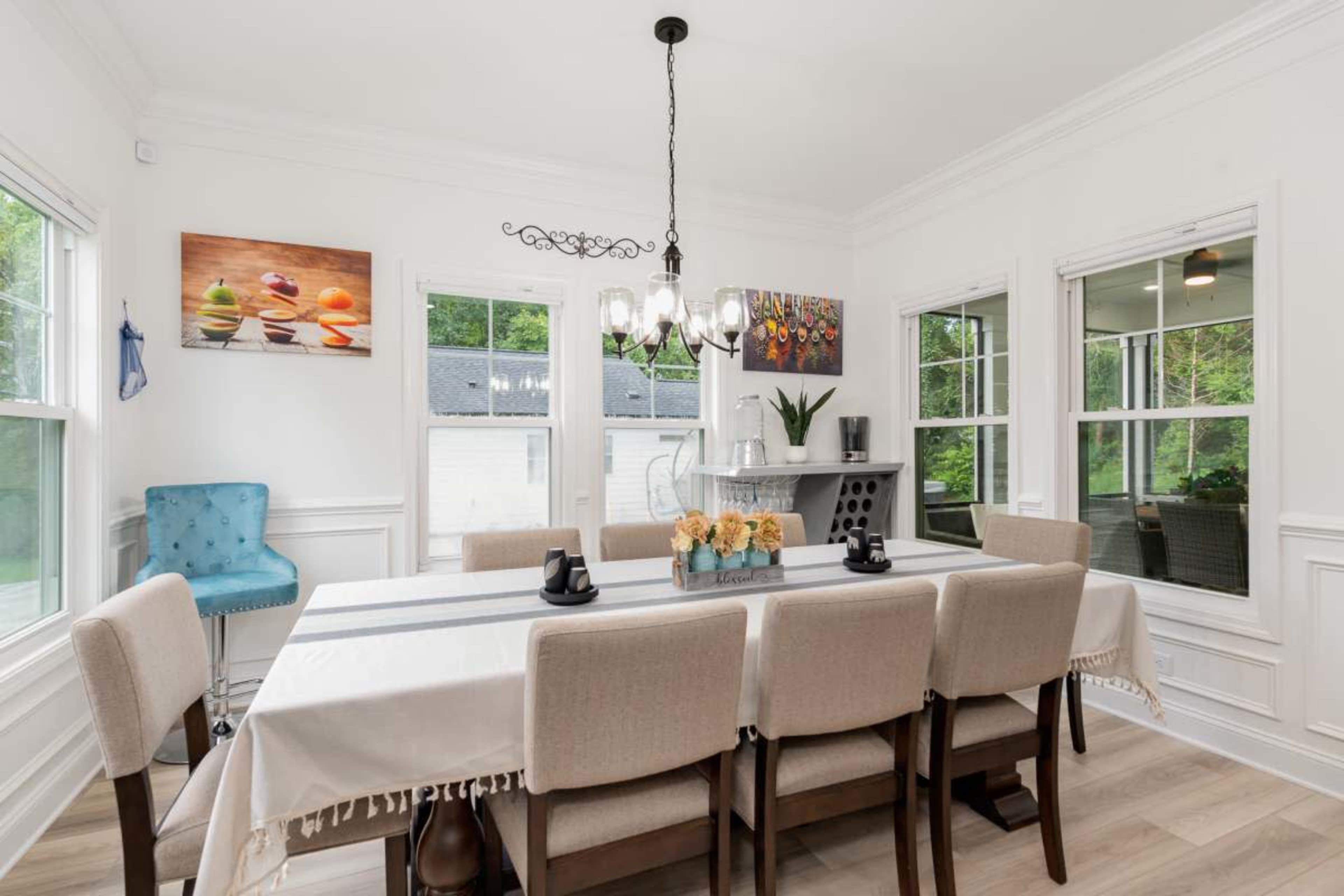 A bright dining room features a long table surrounded by beige chairs, with large windows and a decorative chandelier above.