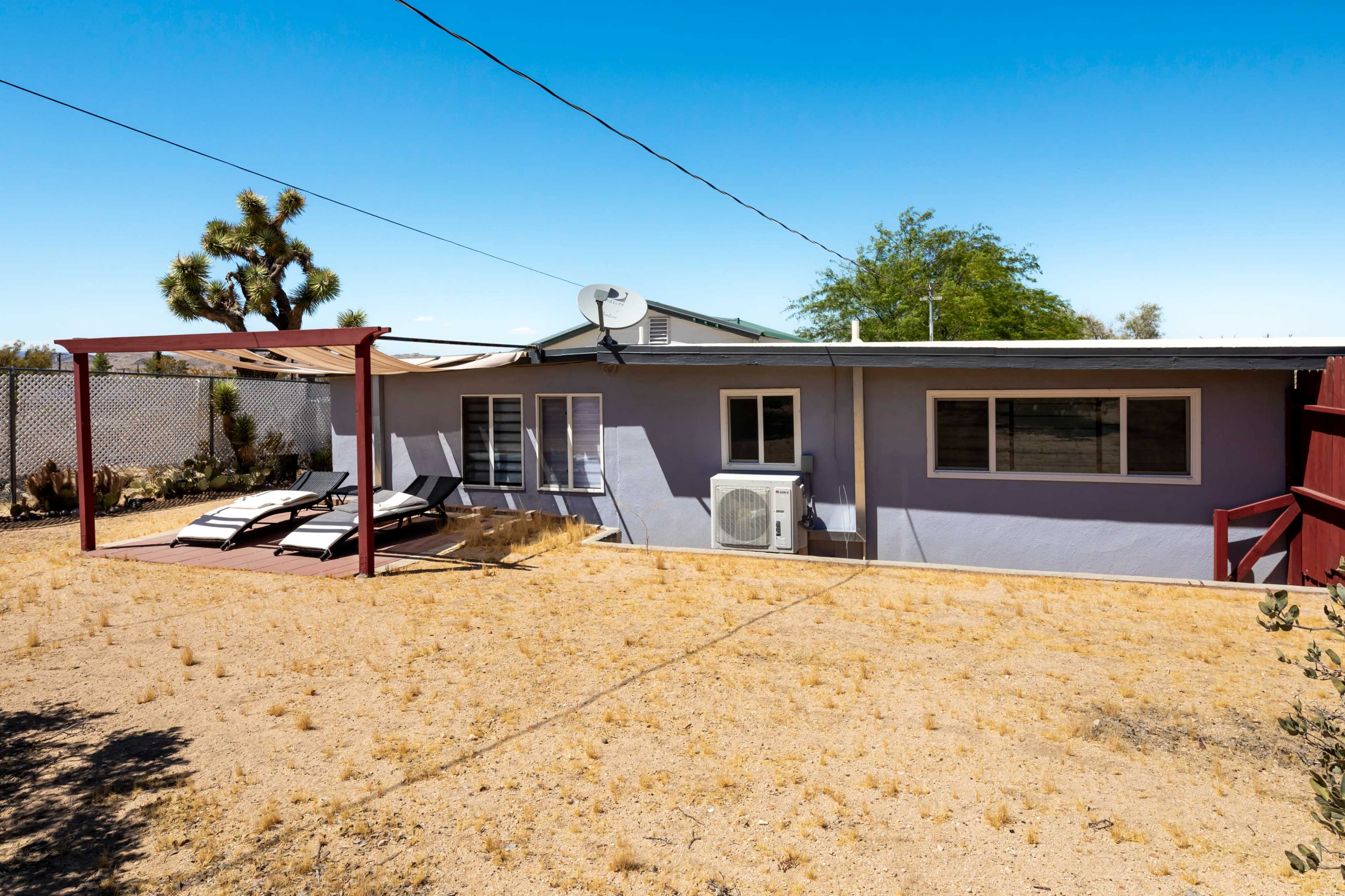 The image shows a simple house with a shaded patio area, two lounge chairs, and an air conditioning unit, set in a dry, arid landscape.