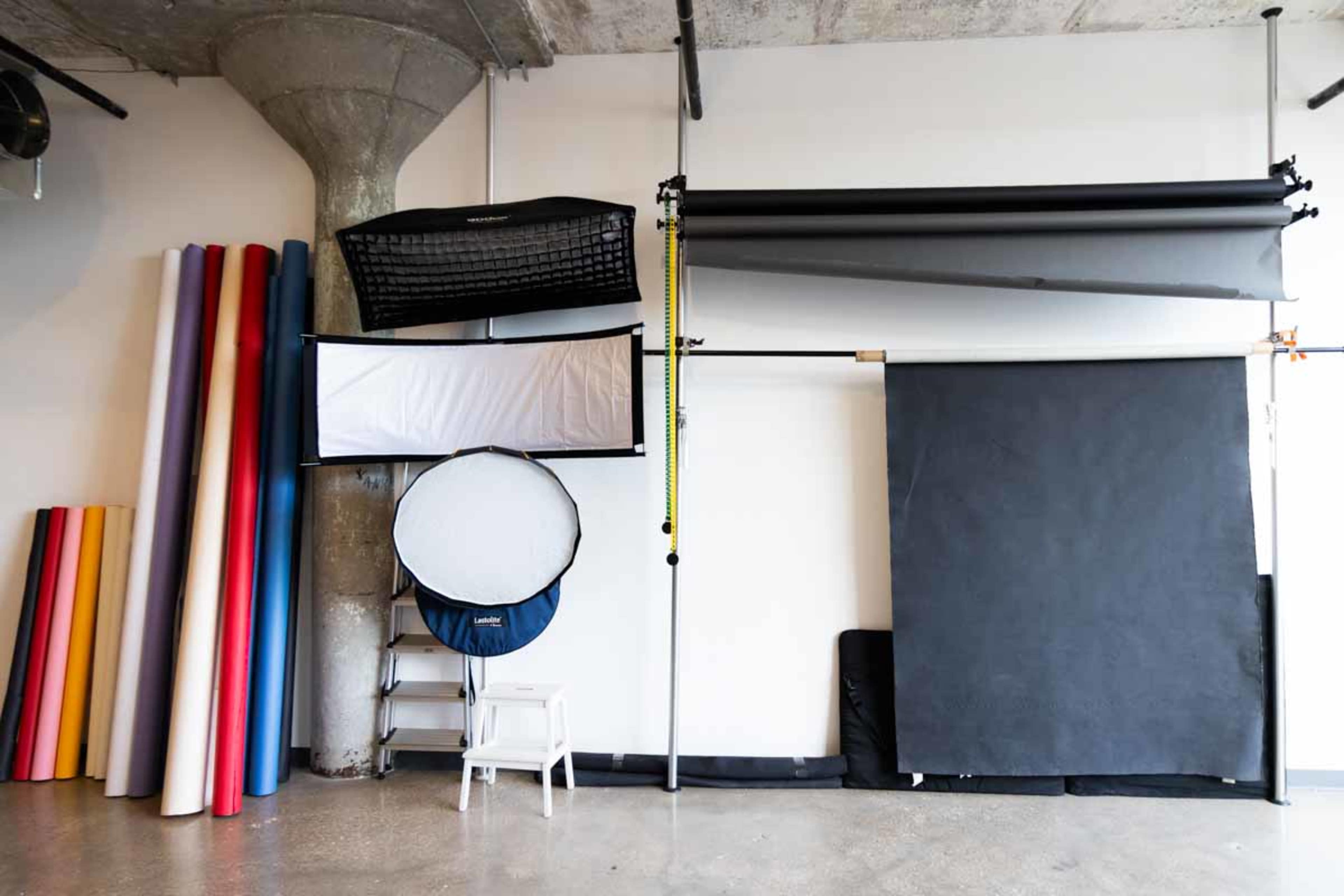 The image shows a photography studio with various backdrops, including large rolls of colored paper and multiple fabric panels, arranged on a wall with a concrete ceiling.