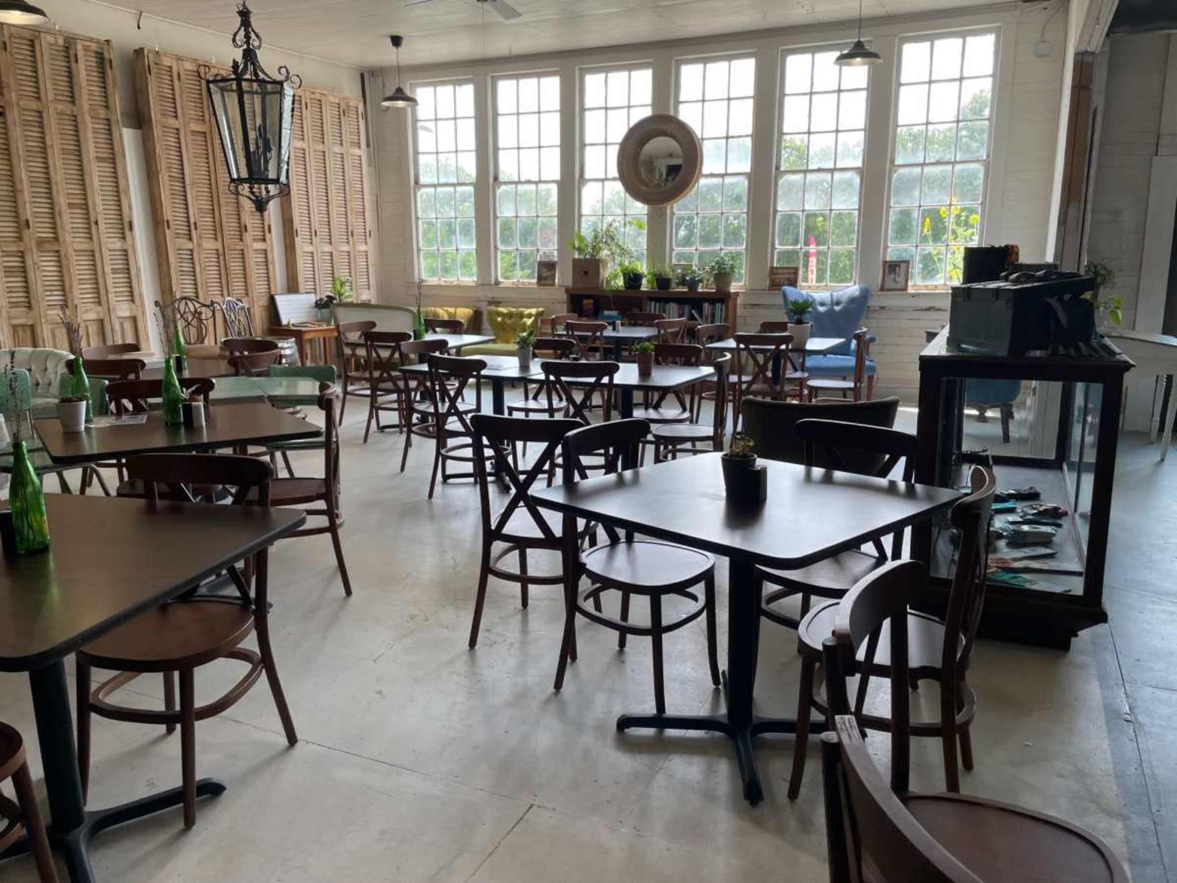 A spacious café with wooden tables and chairs arranged neatly, large windows allowing natural light to enter, and decorative elements like plants and a display case in the background.