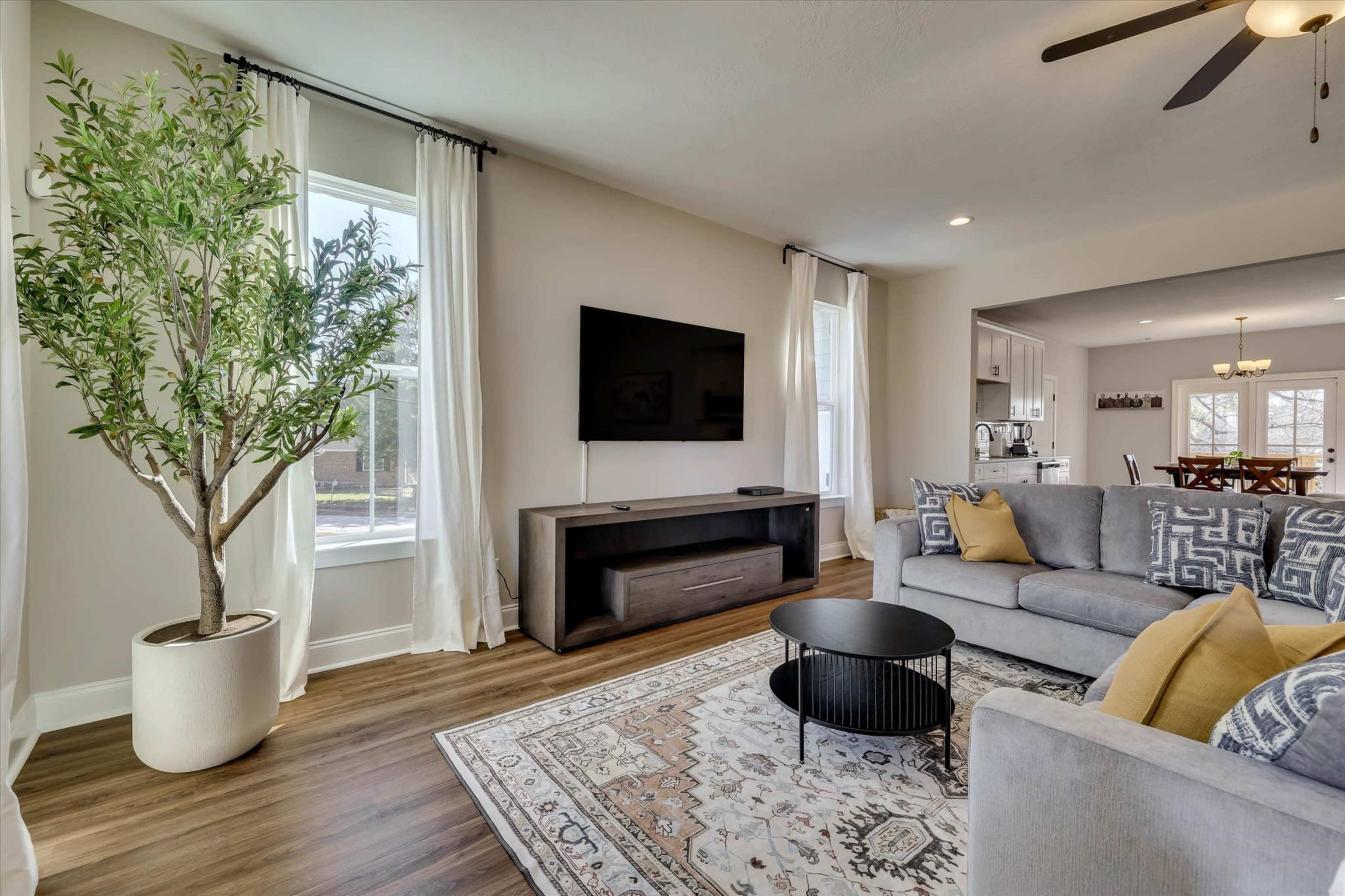 The image shows a modern living room featuring a gray sectional sofa, a round black coffee table, and a large television mounted on the wall, with a potted plant and light curtains by the window.