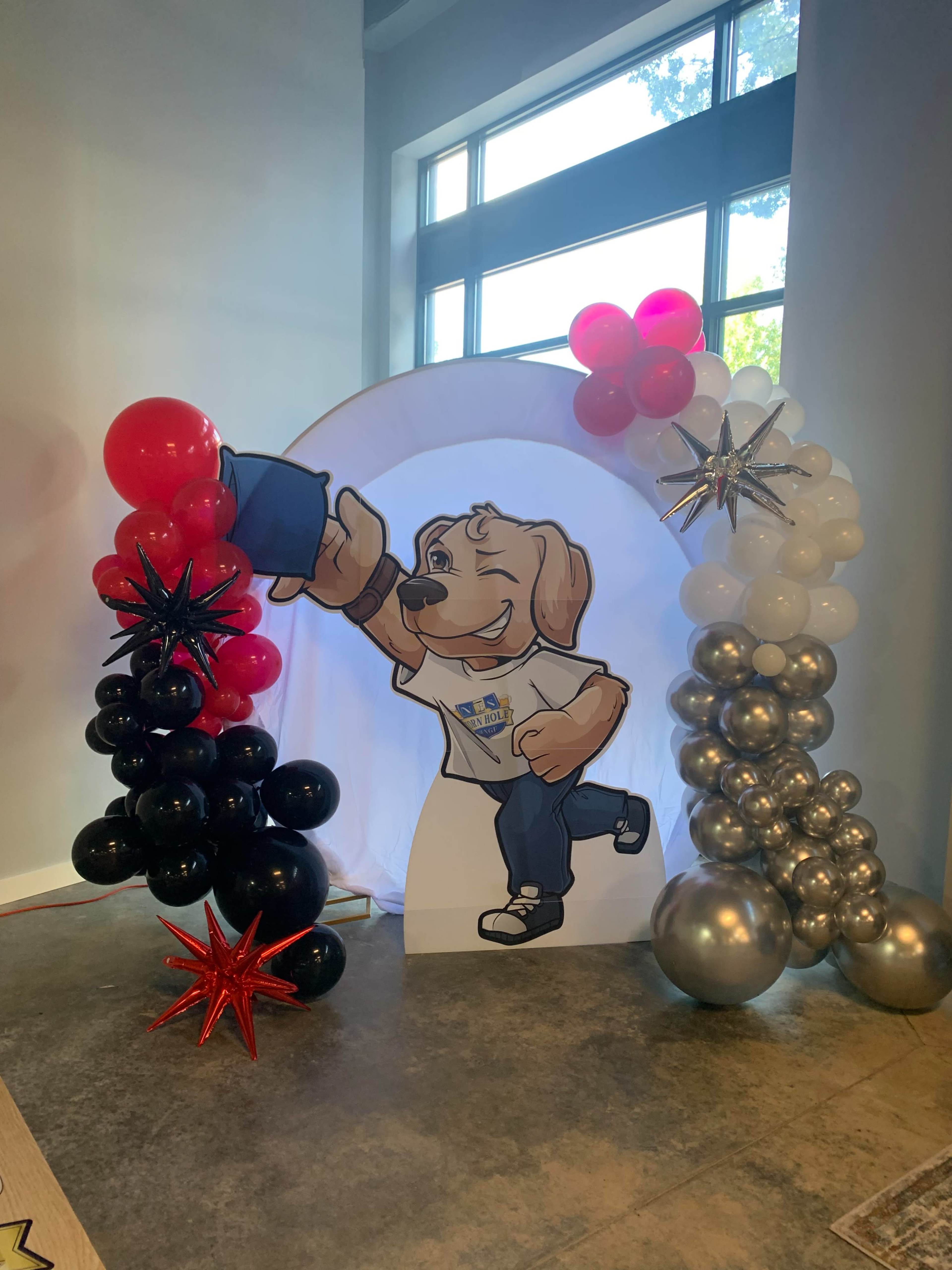 The scene features a balloon arch with black, red, and silver balloons surrounding a cutout of a cartoon dog character holding a blue object.