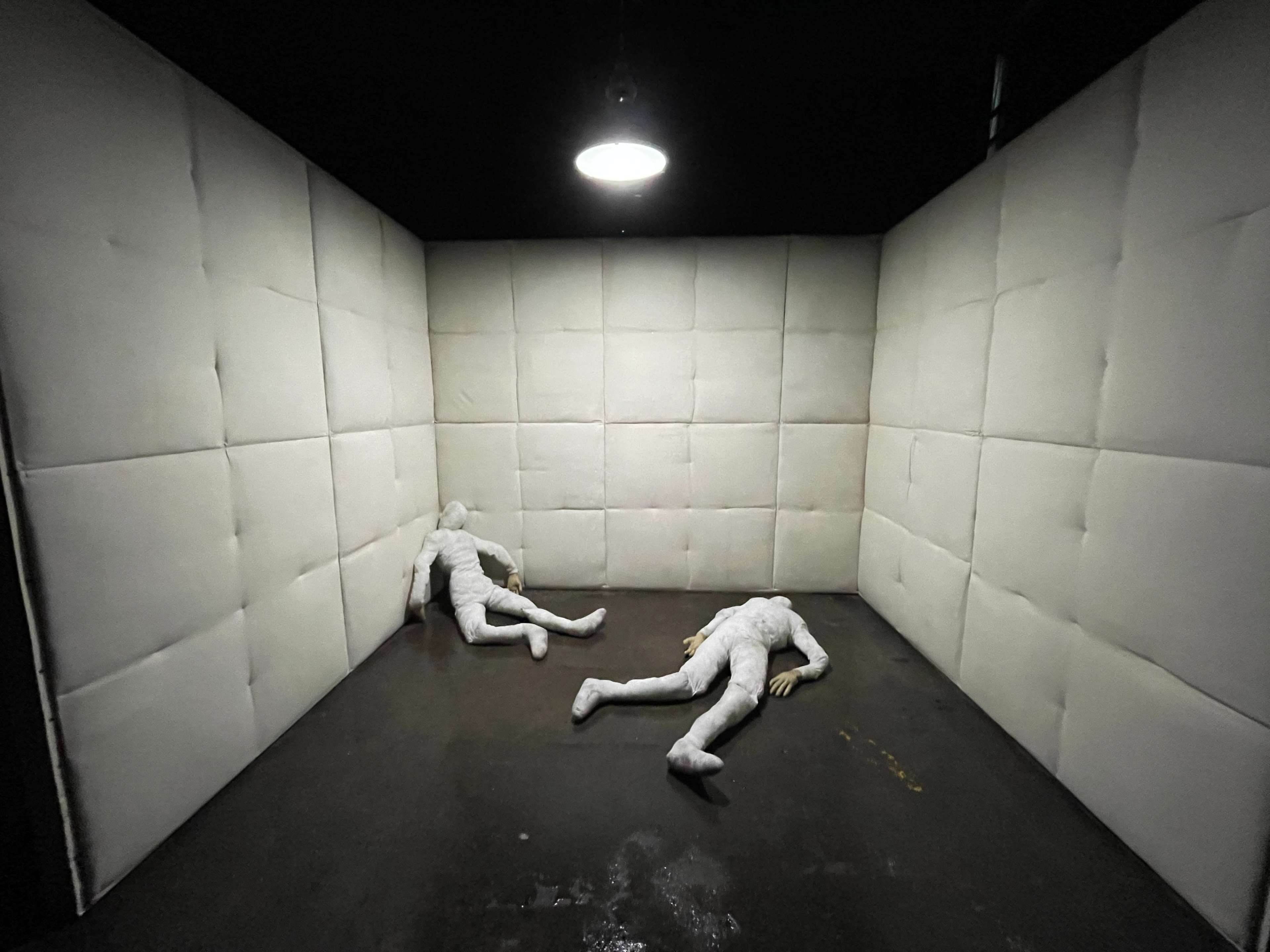 Two life-sized, featureless figures lying on the floor of a small, padded room with white walls.