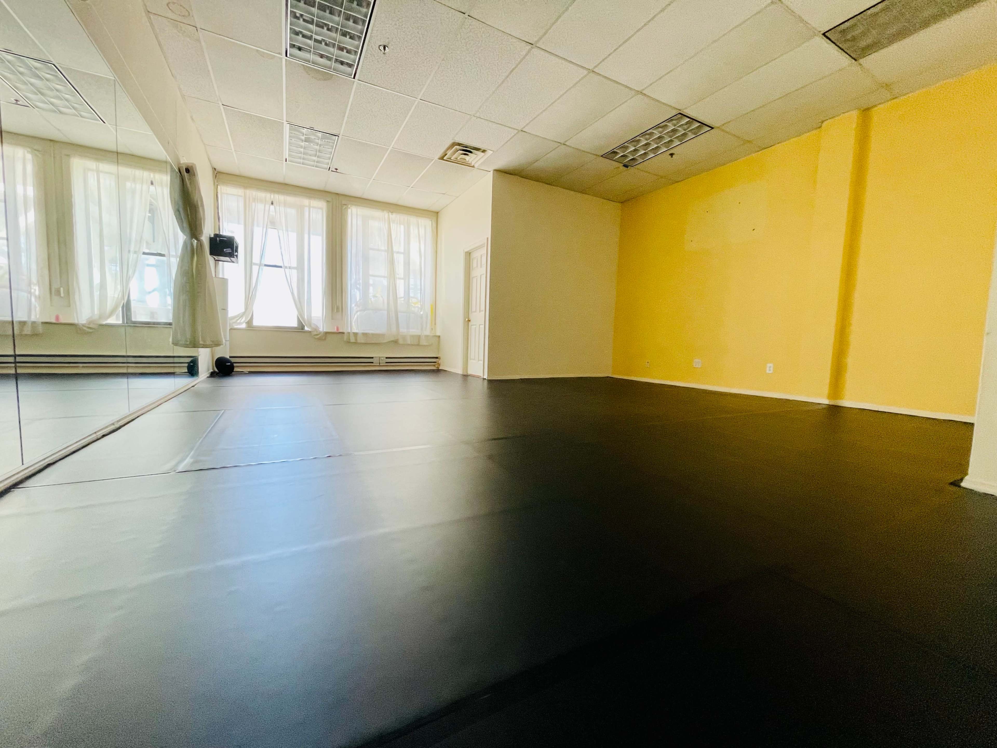 An empty room with black flooring, large windows, and a mirror along one wall.