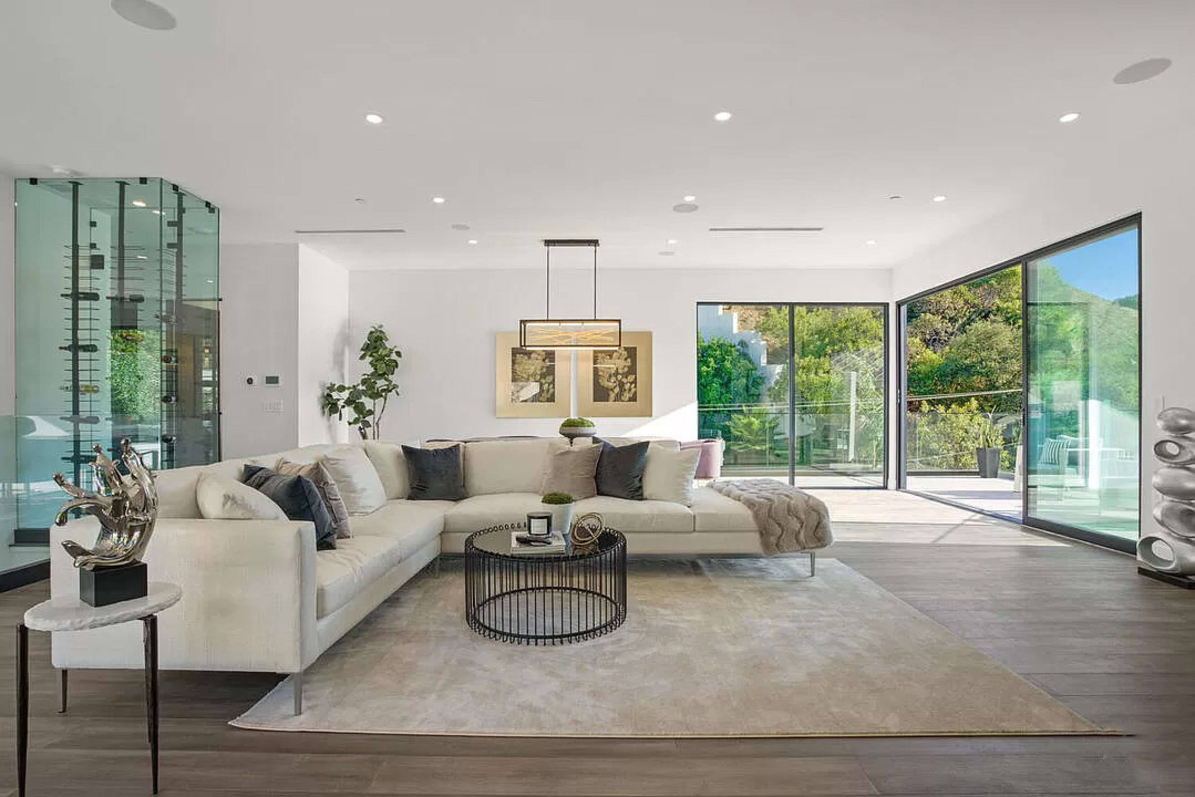 The image shows a modern living room featuring a large sectional sofa, a round black coffee table, and floor-to-ceiling windows that open to a green outdoor area.