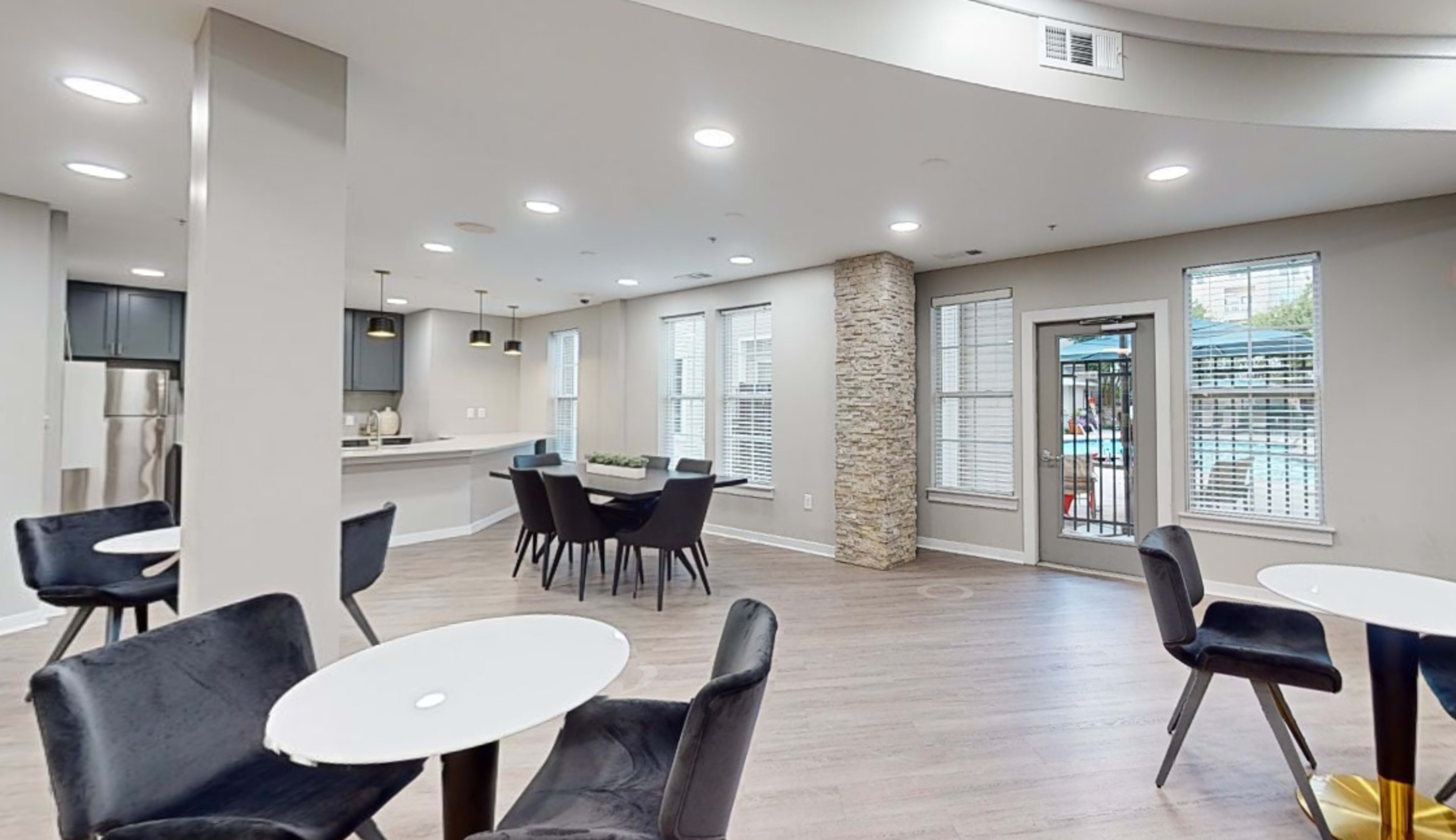 The image shows a modern common area with a kitchenette and seating arrangement, featuring a view of a pool area through large windows.