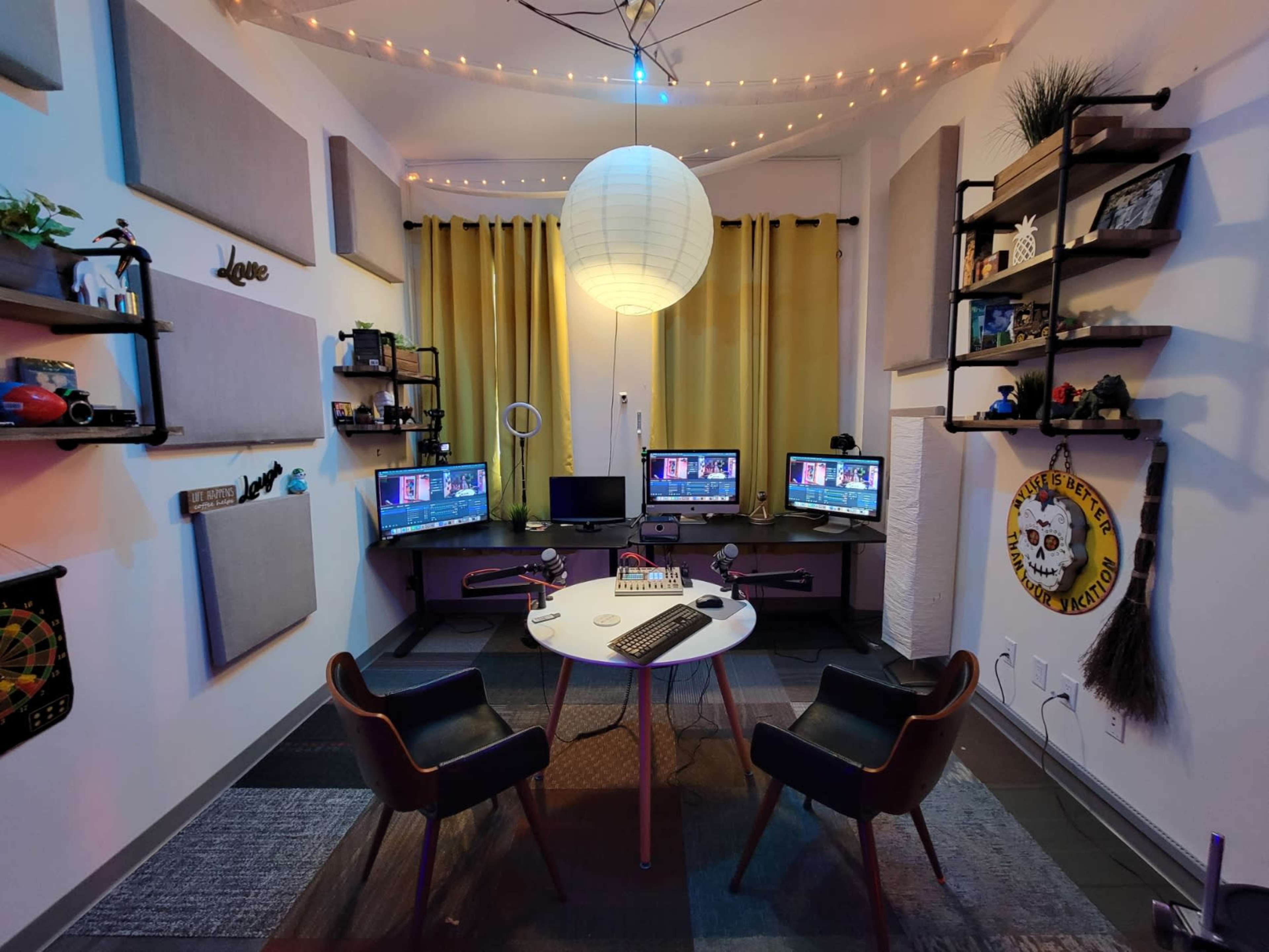 A dual workstation setup with two computer monitors, a circular table, and two chairs in a room decorated with shelves, plants, and soft lighting.