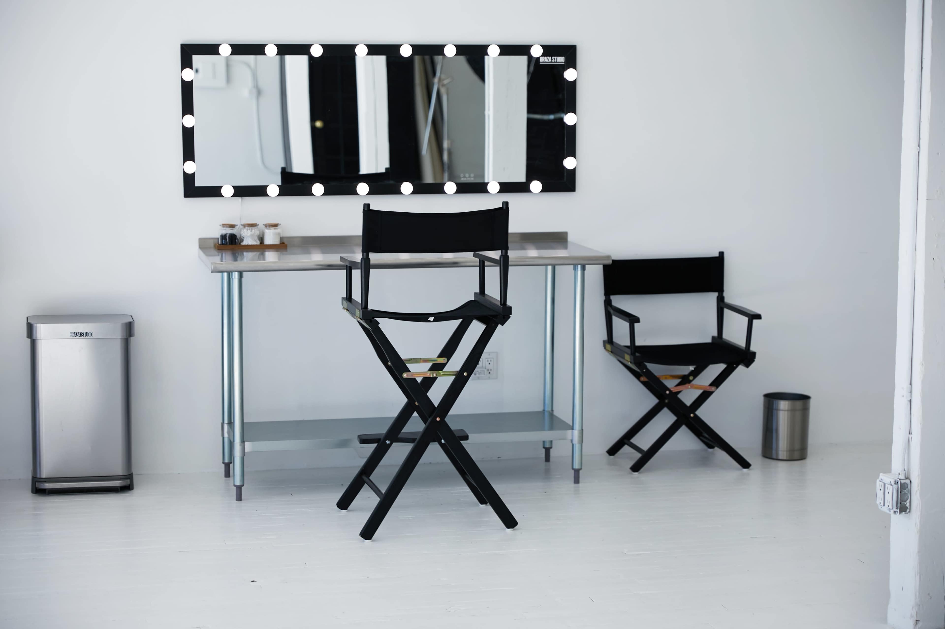 A bright, minimalist dressing room features two black director's chairs, a stainless steel table with lights above a mirror, and a trash bin in the corner.