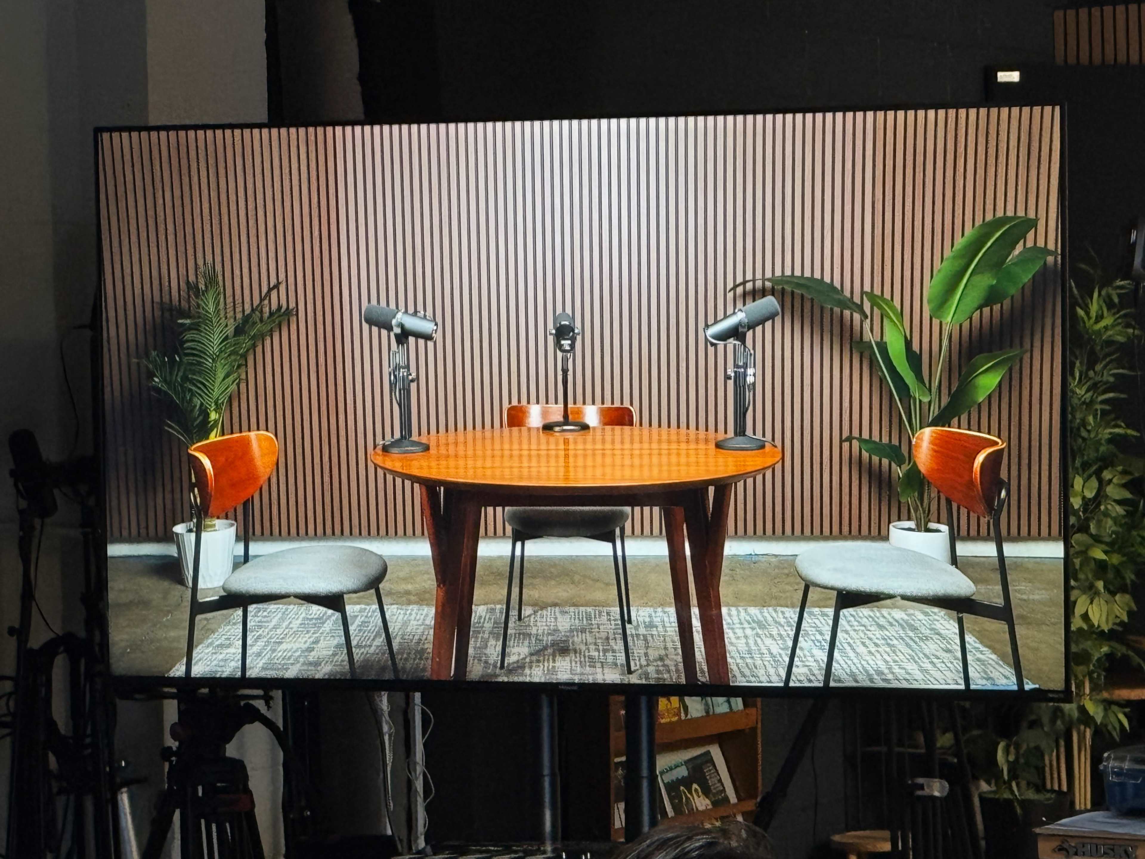 The image shows an empty podcast studio with a round table, three microphones, and two chairs facing each other, surrounded by decorative plants against a wooden panel backdrop.