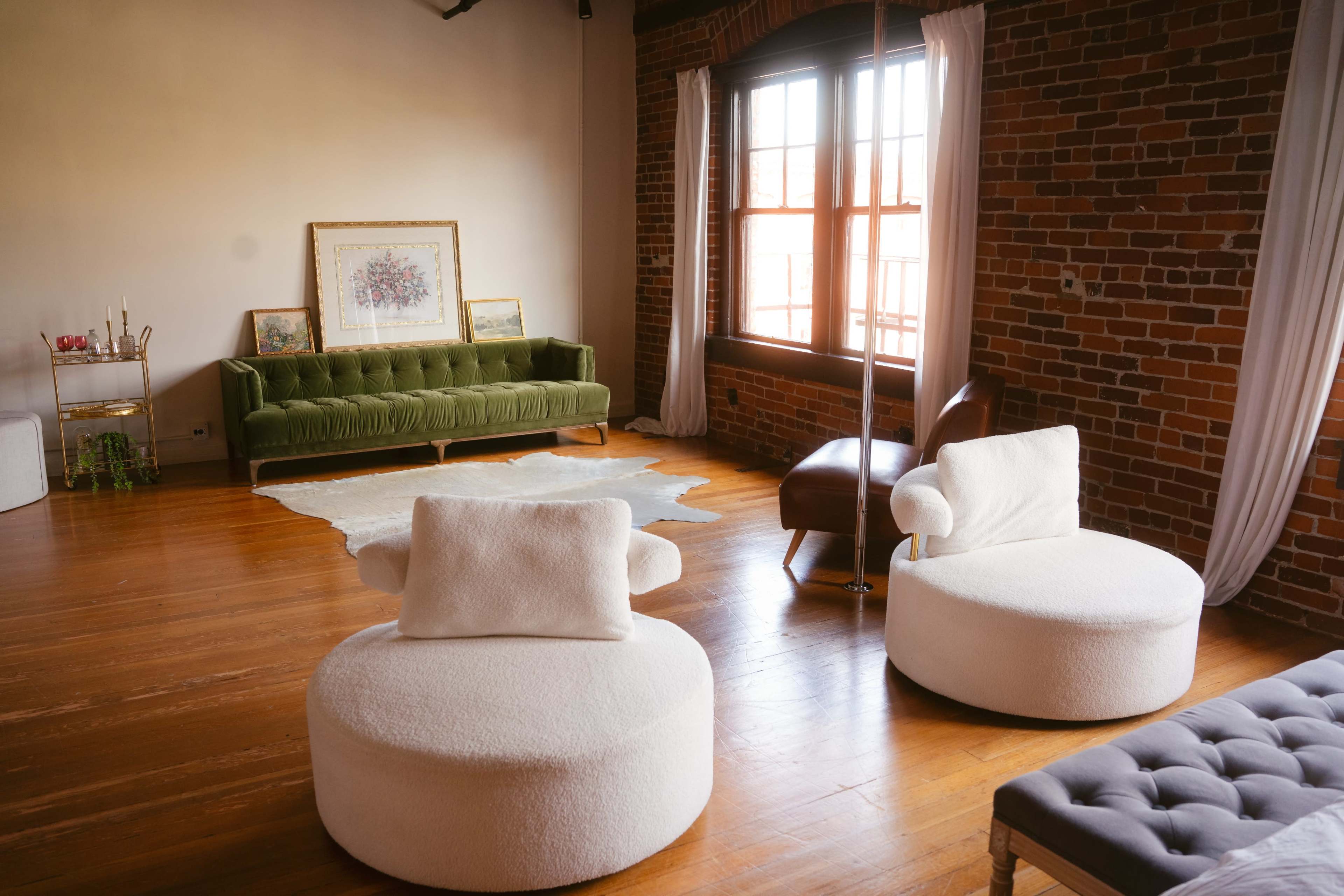 The image shows a spacious room with exposed brick walls, featuring a green velvet sofa, two white circular chairs, and a brown leather chair near a window.