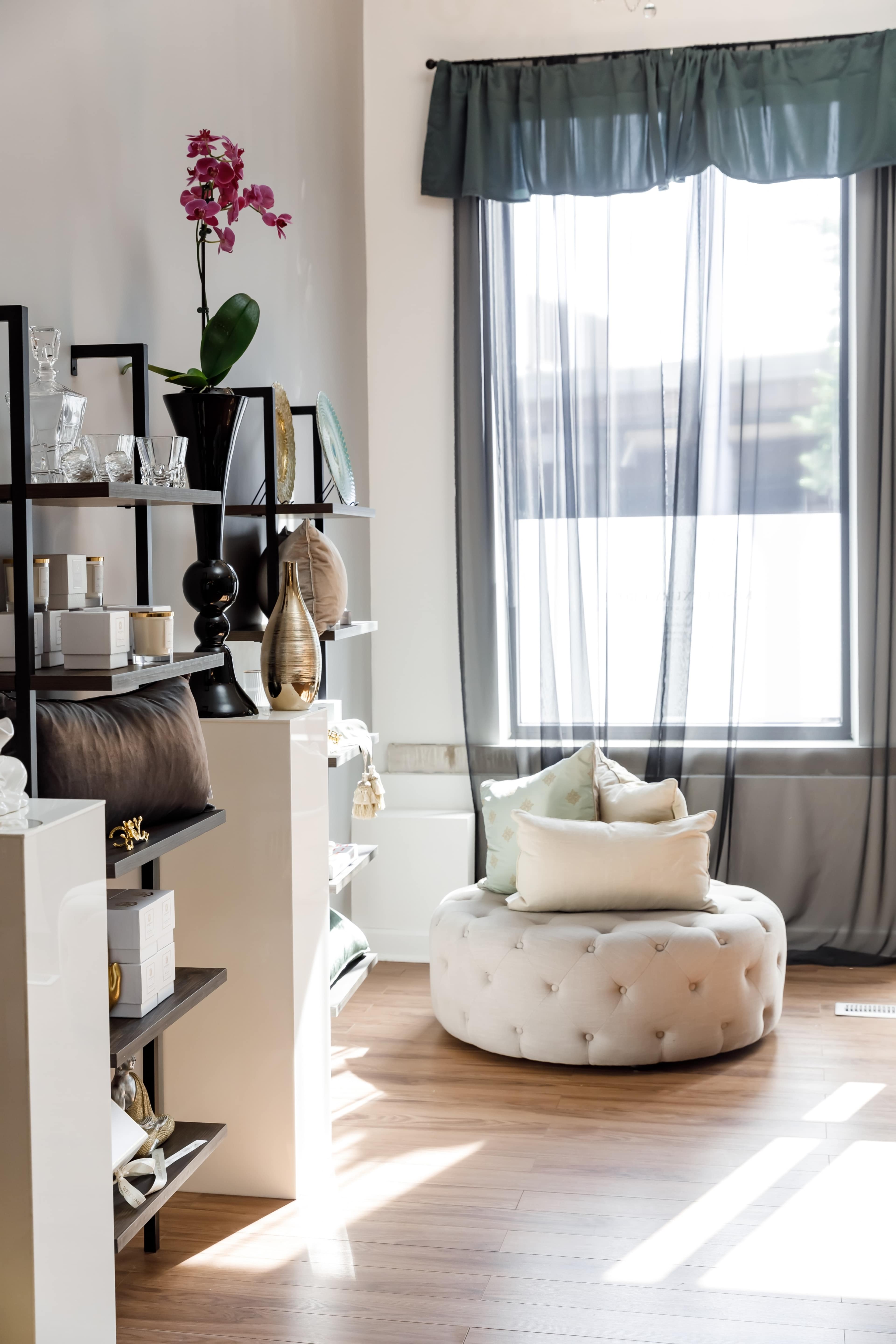 A bright room with a round tufted ottoman and shelves displaying various decorative items, complemented by a large window with sheer curtains.