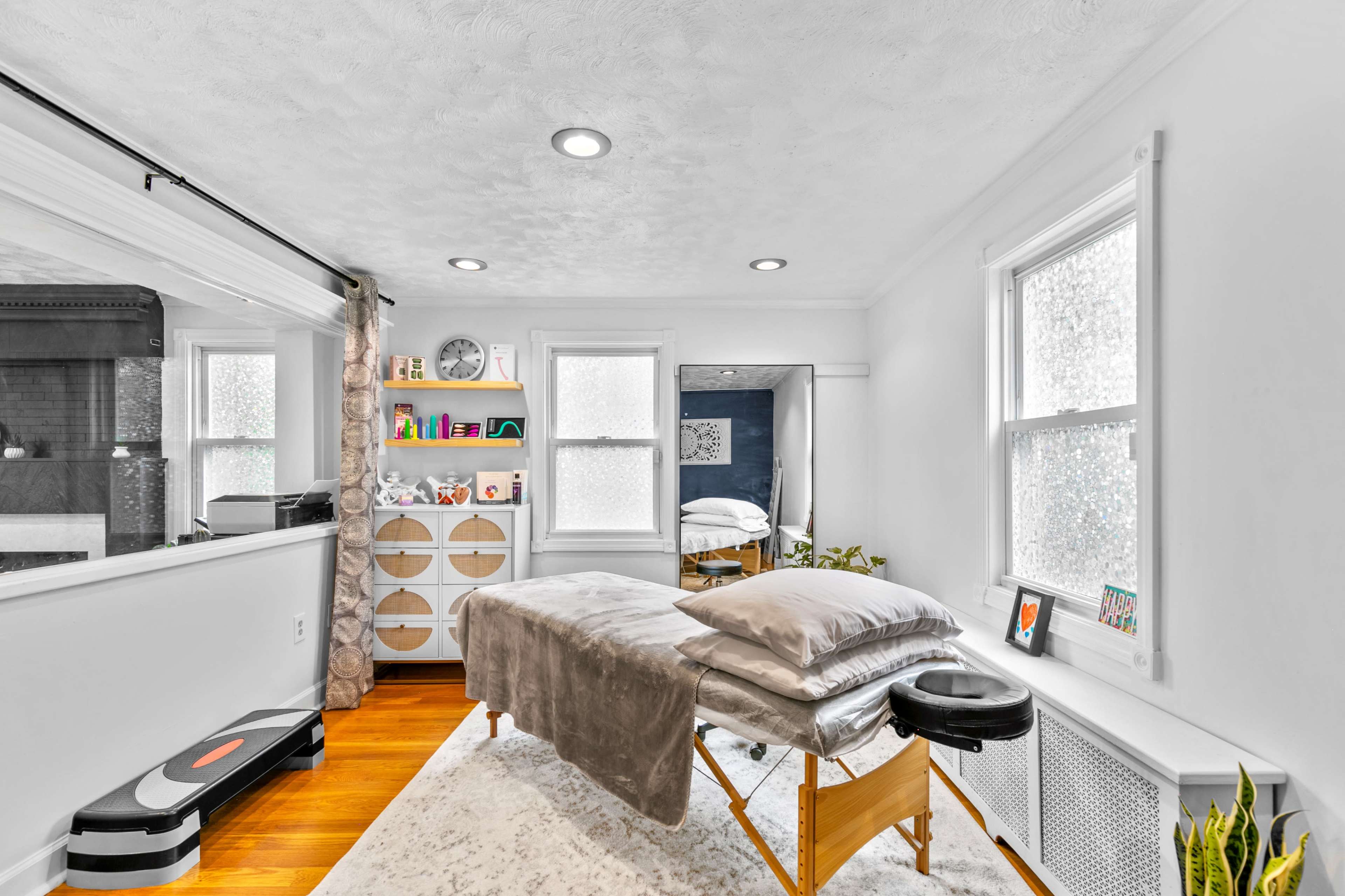 The image shows a bright and modern therapy room with a massage table, shelves displaying decor, and large windows allowing natural light.