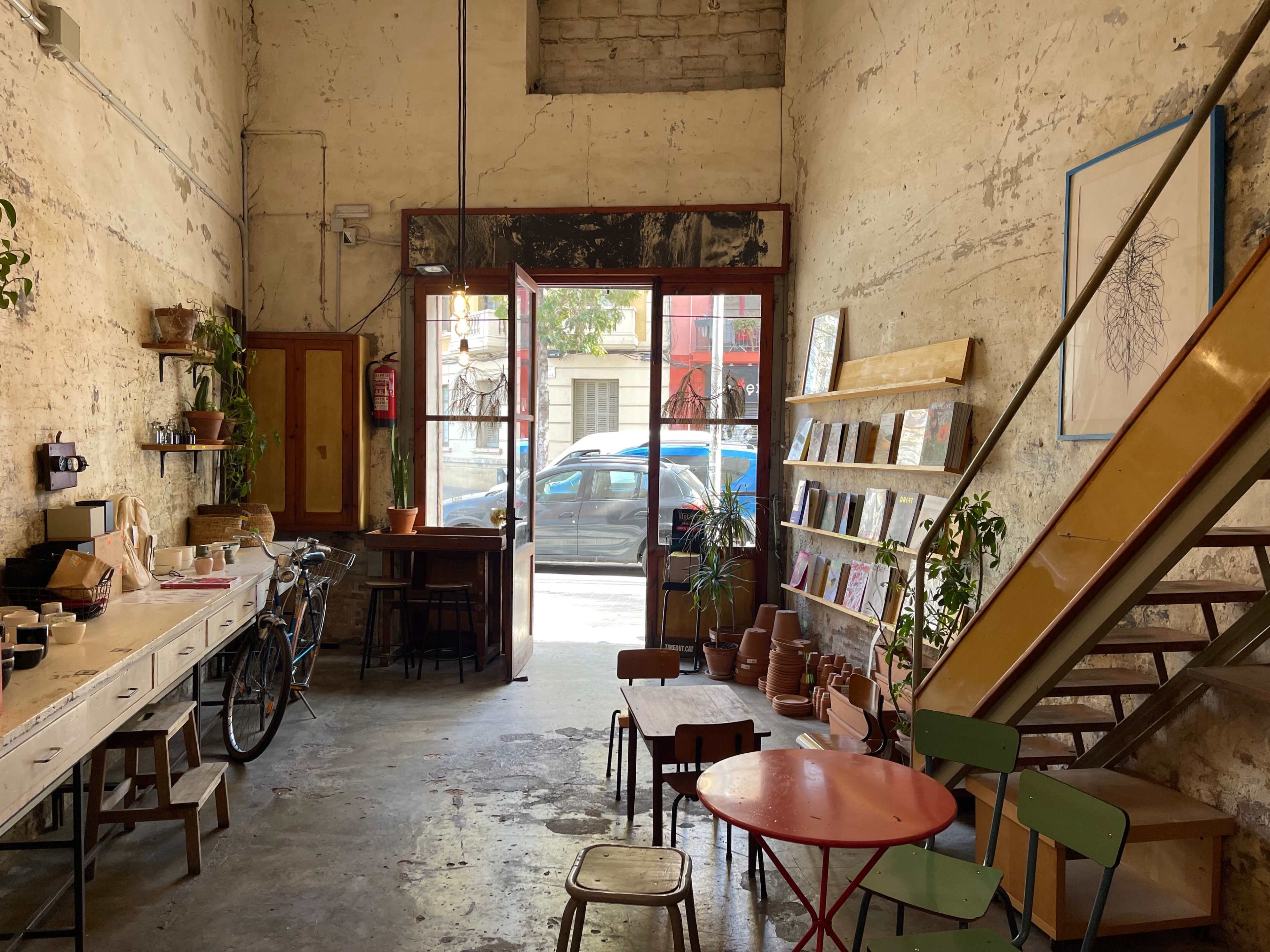 A spacious interior of a café features a wooden counter, a staircase, a seating area with colorful chairs, and large windows that open to the street.