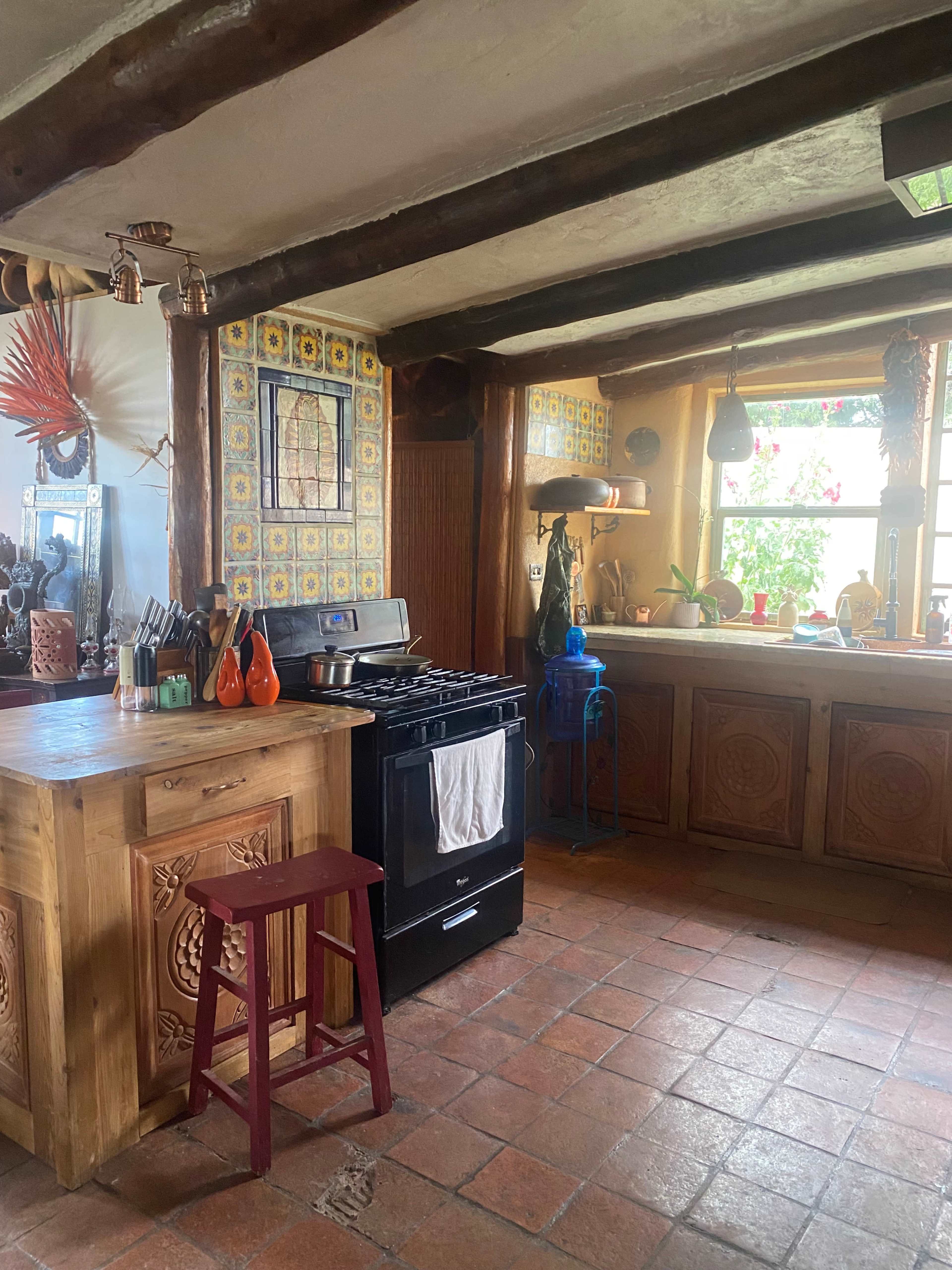 The kitchen features a wooden island, a black stove, and colorful tiled walls with a window overlooking a garden.
