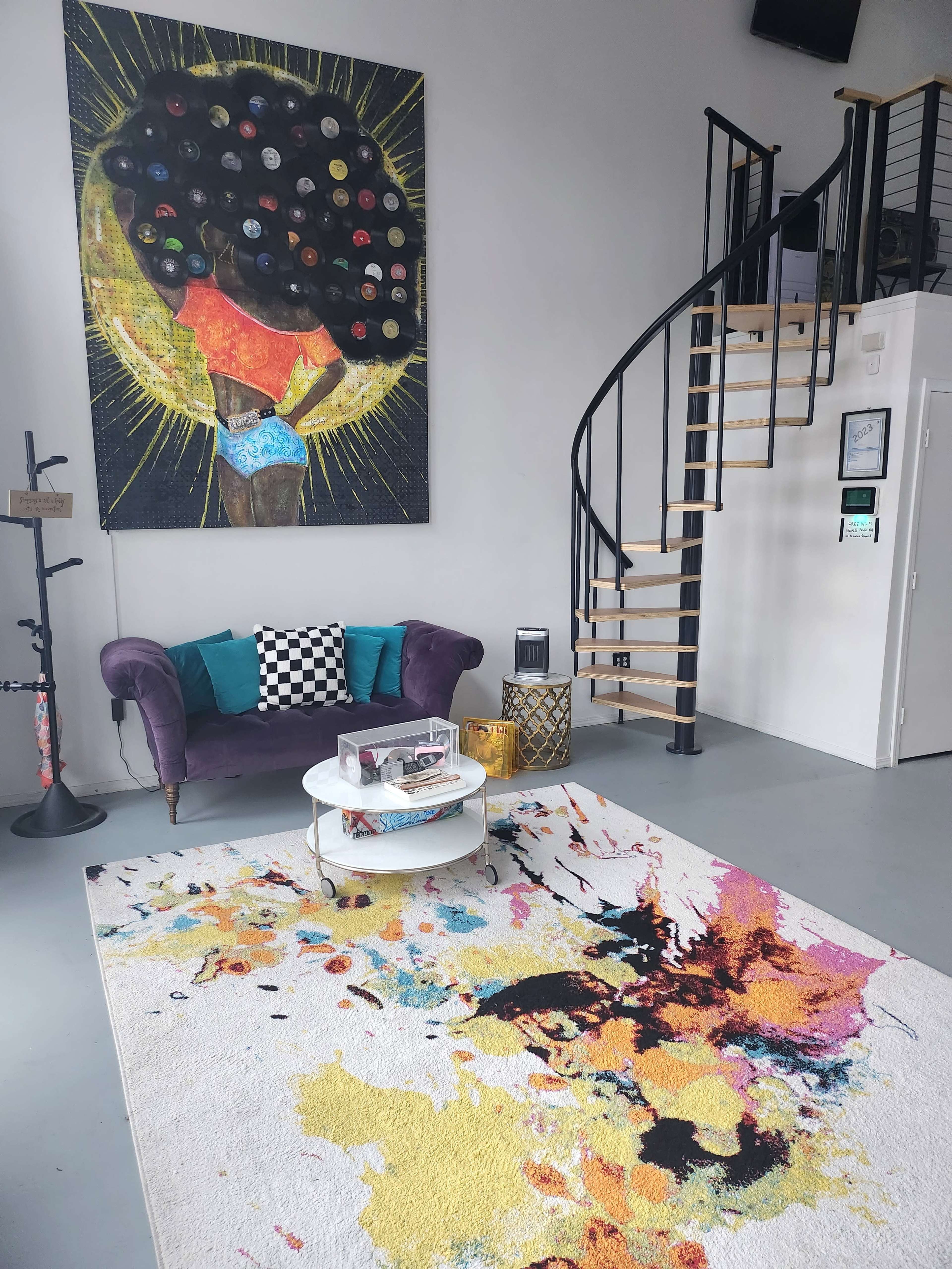 The room features a spiral staircase, a purple couch, a colorful area rug, and a large wall painting of a figure with an elaborate tree hairstyle.