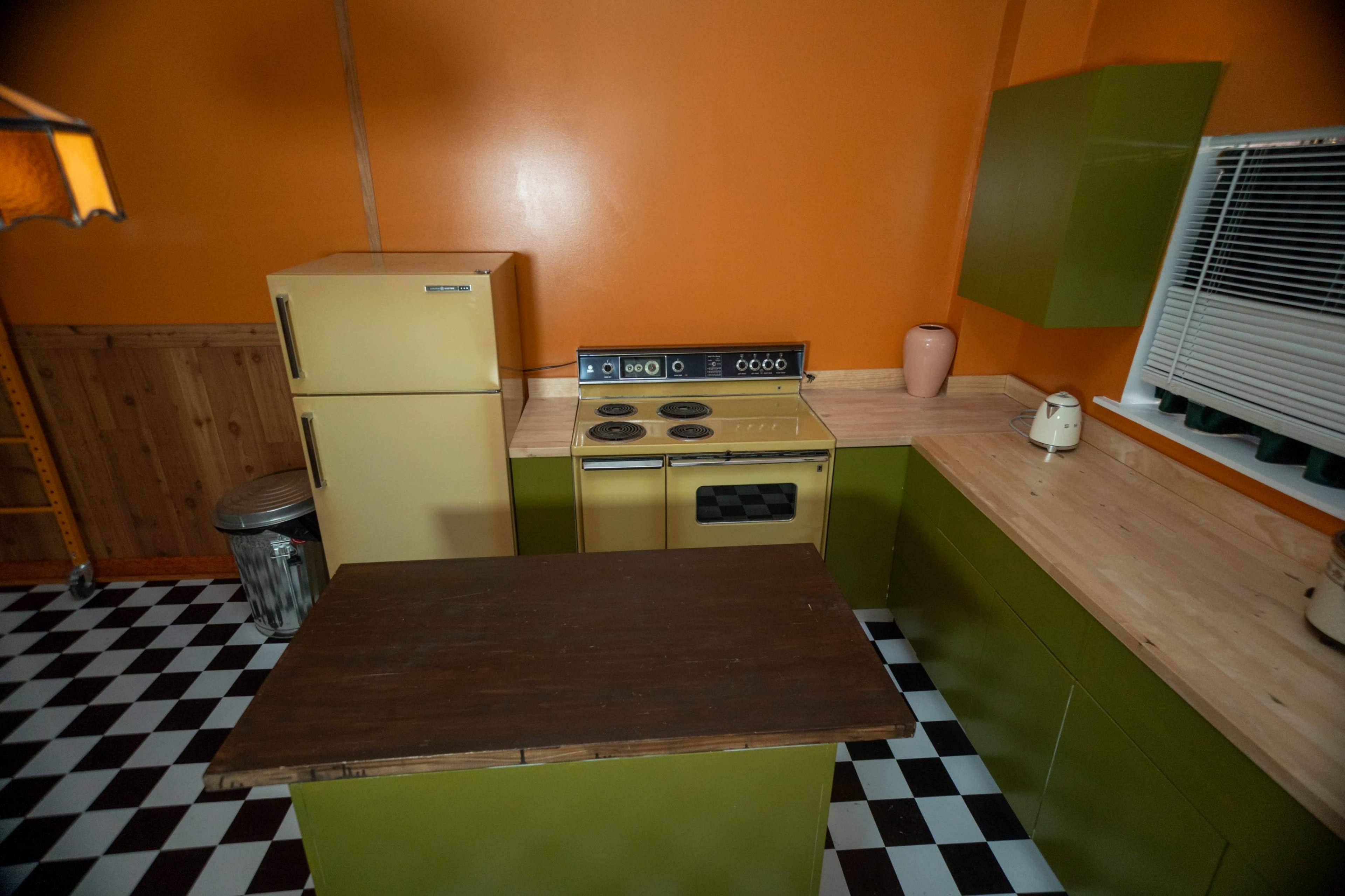 The image shows a kitchen with orange walls, green cabinetry, a yellow refrigerator, and a checkered black-and-white floor.
