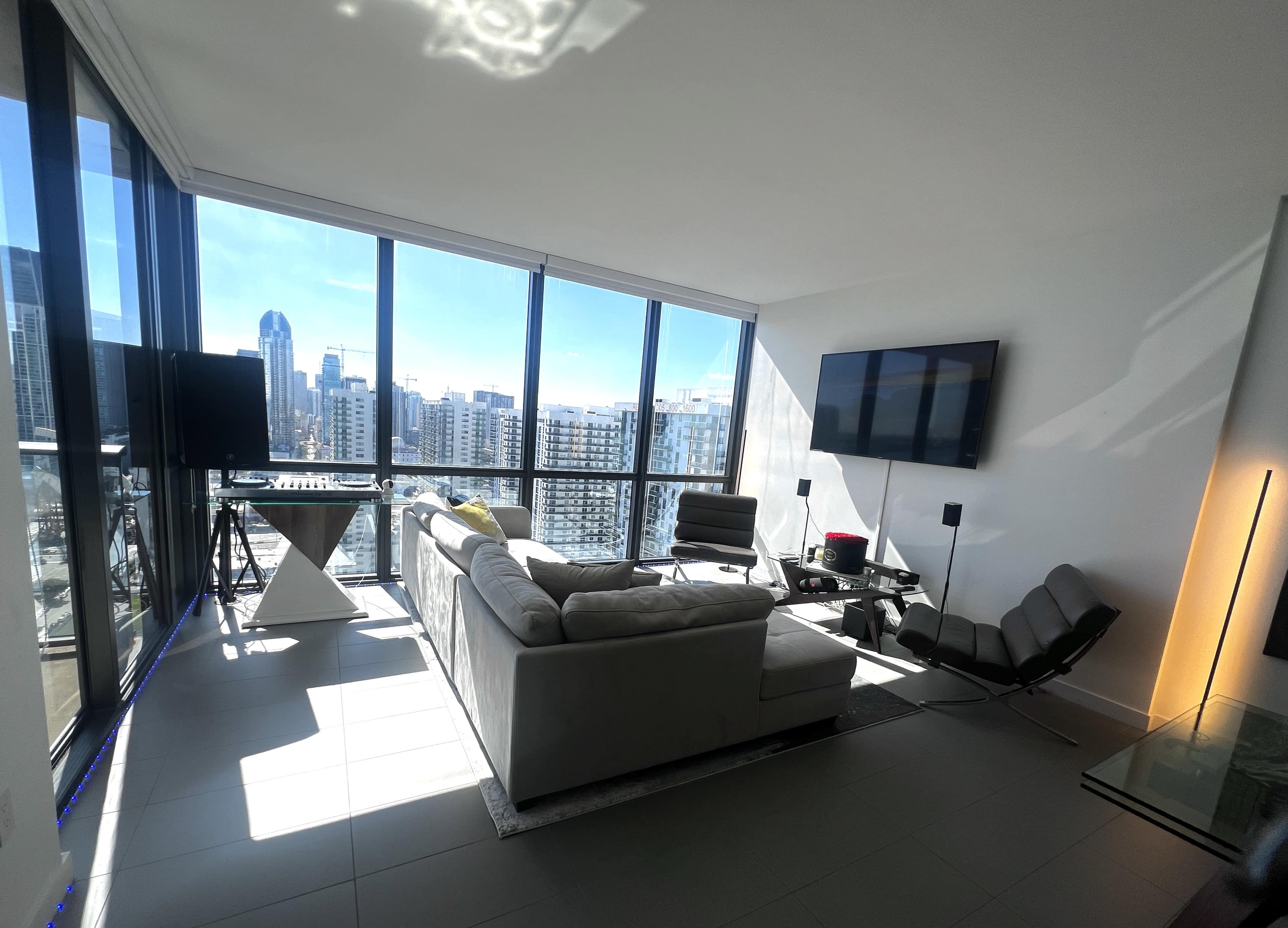 A modern living room features a large window with a city view, a sectional sofa, a sleek television, and minimalist furniture.