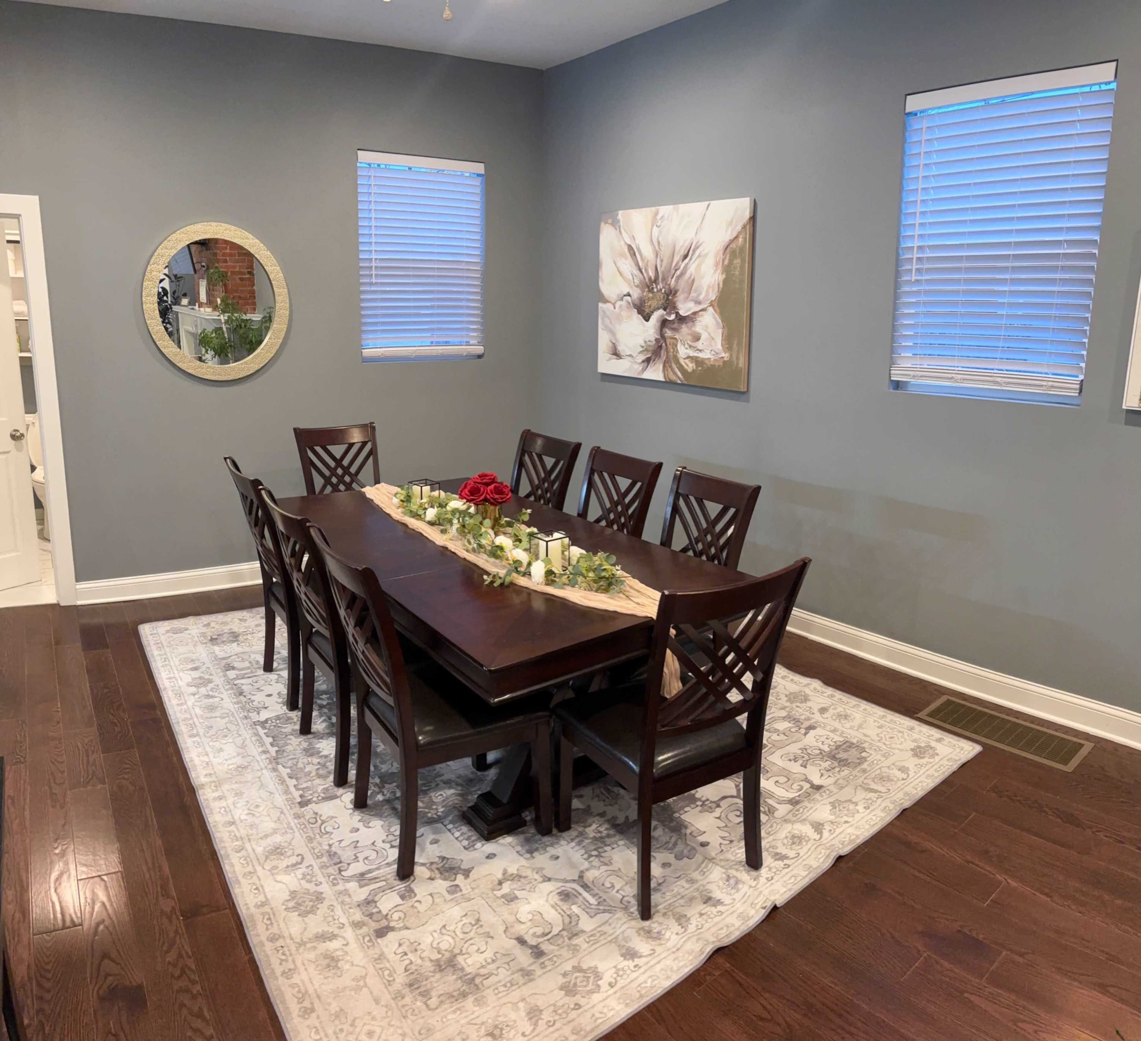 A rectangular dark wood dining table with eight chairs is set on a patterned rug in a room with gray walls and two windows.