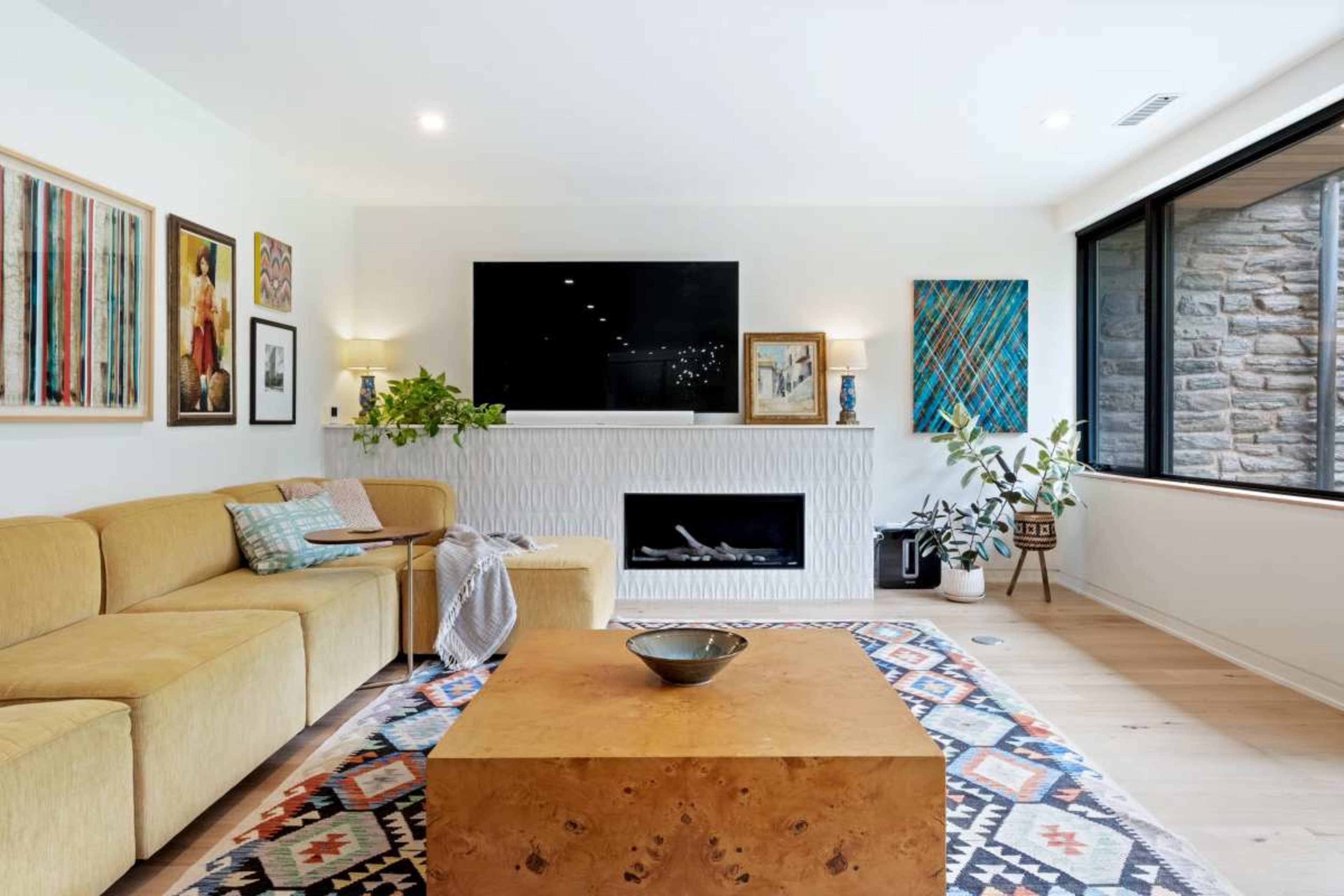 A modern living room features a yellow sectional sofa, a large flat-screen TV above a white fireplace, and a decorative wooden coffee table on a patterned rug.