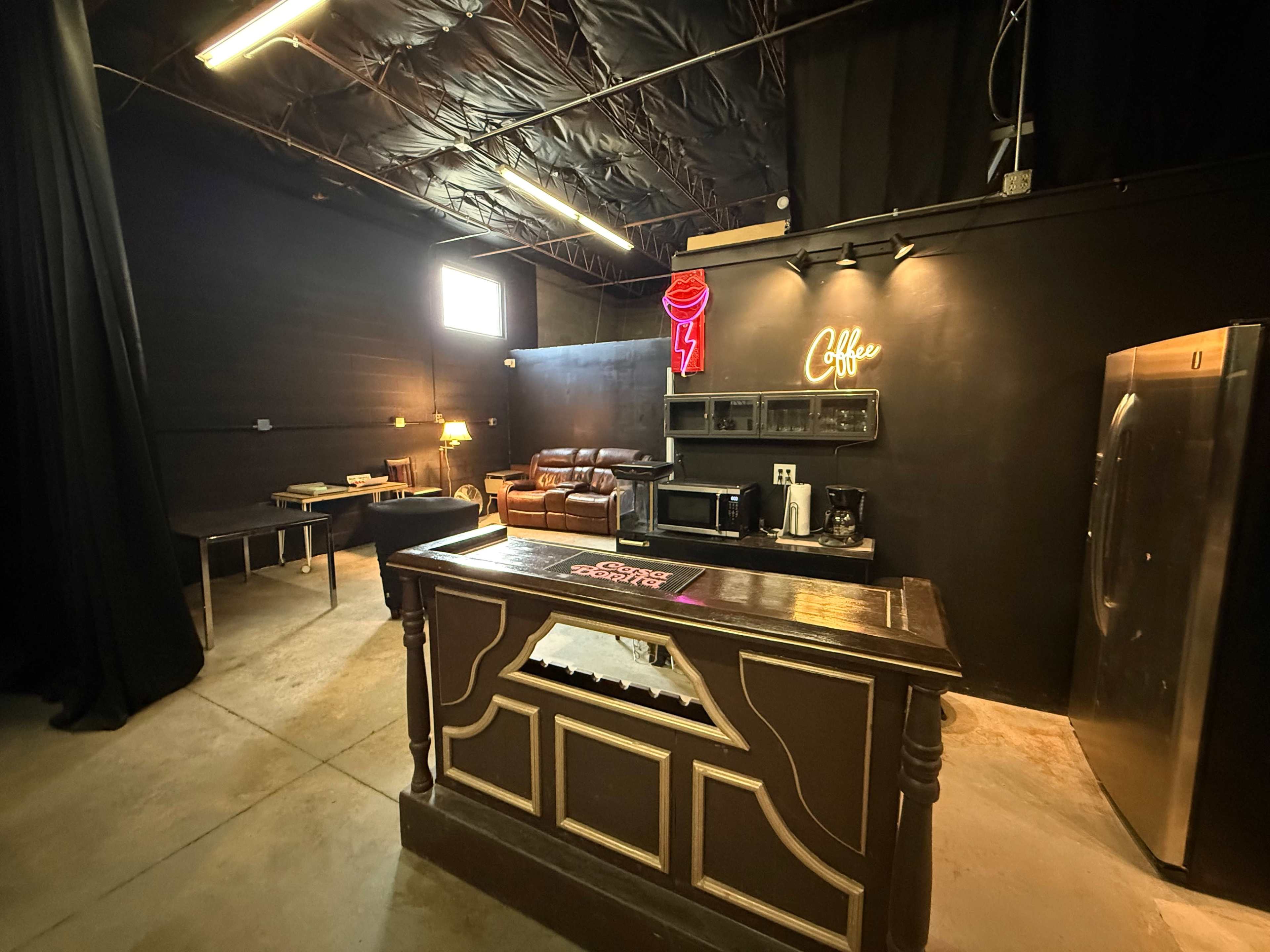 The image shows an industrial-style coffee shop interior with a wooden counter, a lounge area featuring a brown sofa, and a kitchenette equipped with a microwave and coffee maker.