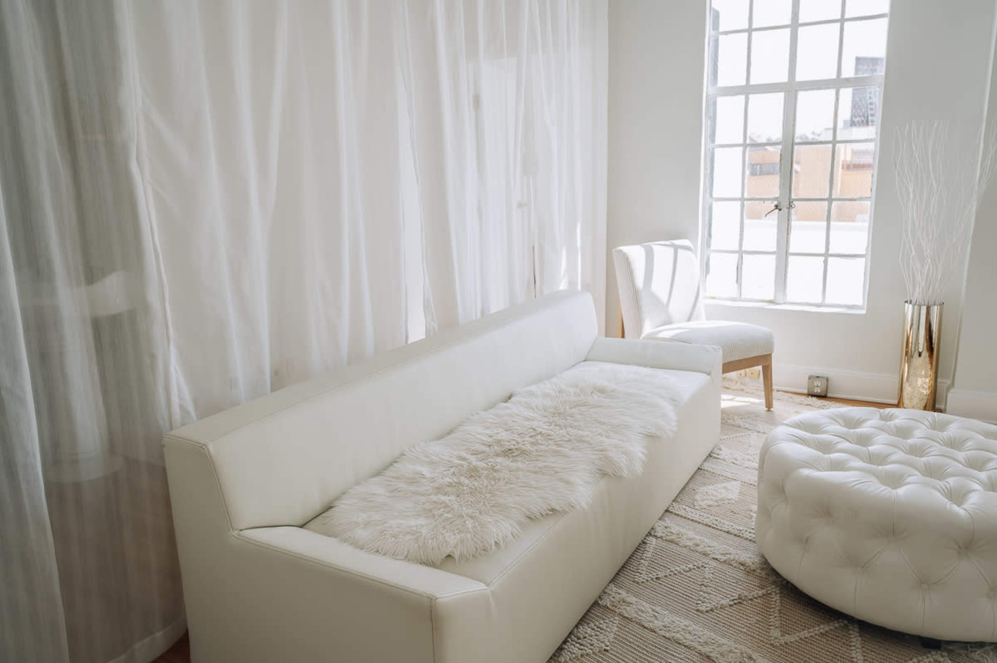 A white sofa with a fur throw is placed next to a window draped with sheer curtains in a light-filled room.