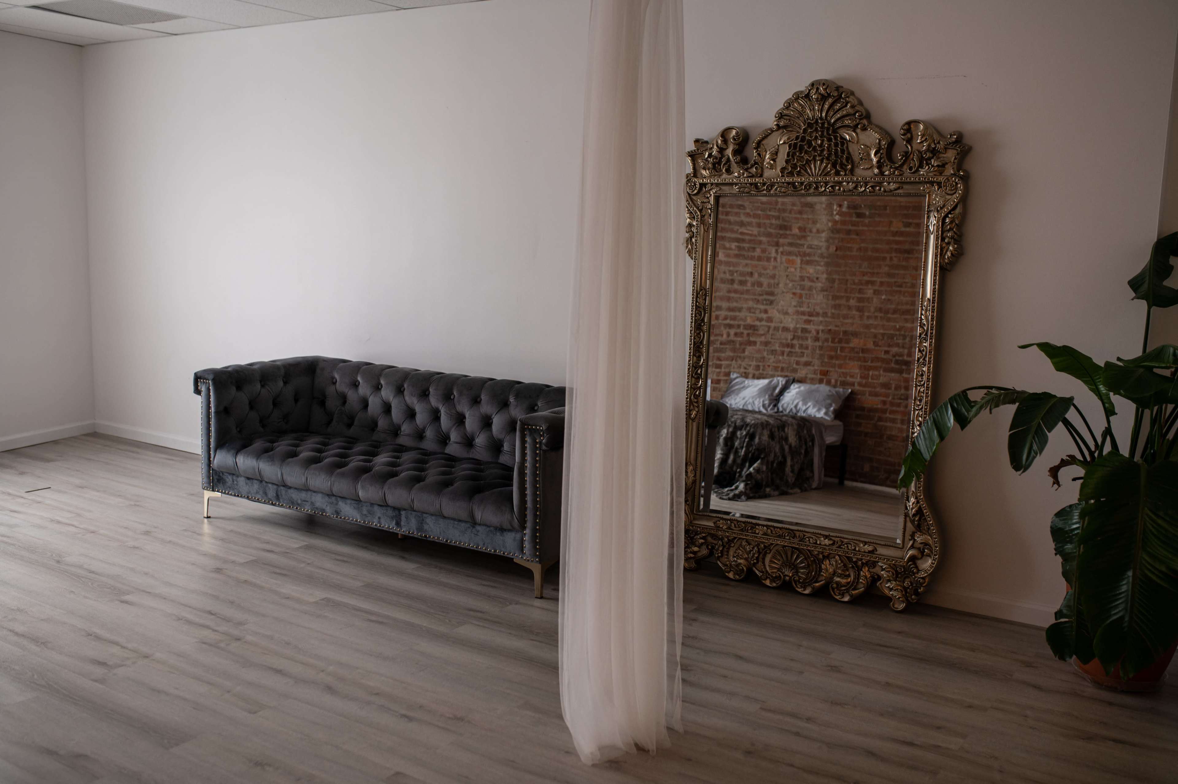 A gray tufted couch facing a large ornate mirror against a brick wall in a minimally decorated room.
