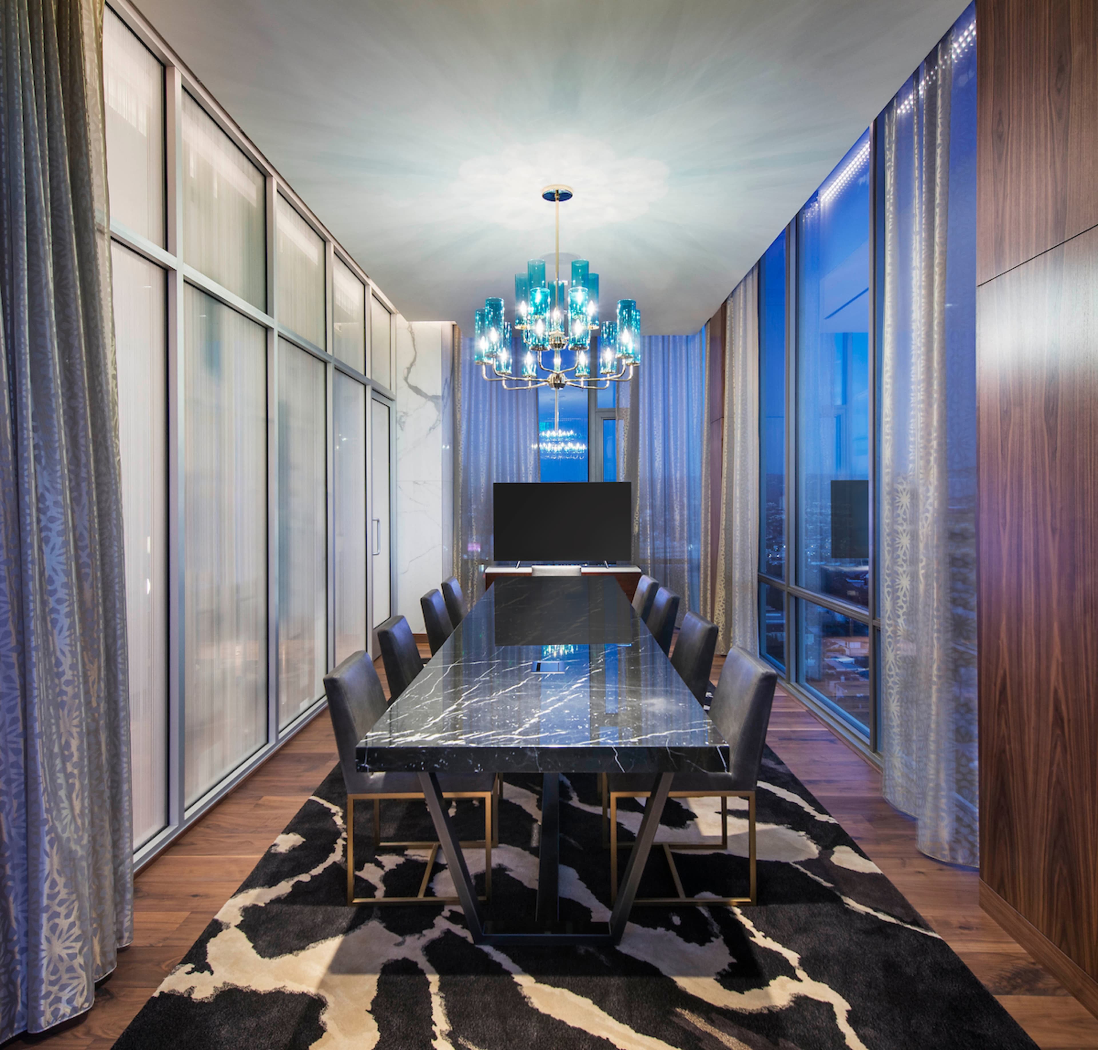 A modern dining room features a long marble table surrounded by black chairs, with a chandelier above and large windows displaying a cityscape at dusk.
